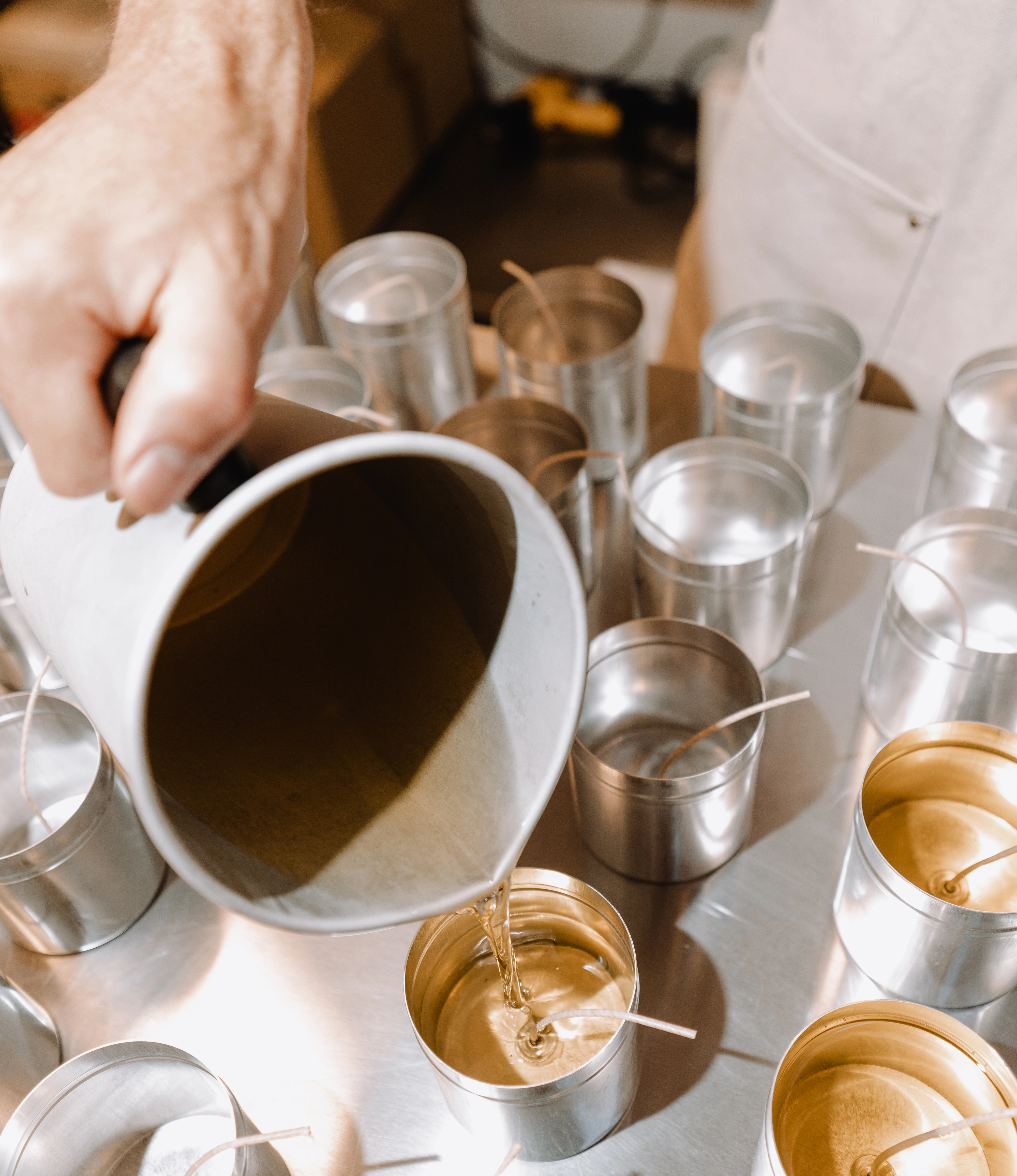 A hand pouring wax into a metallic candle tin, surrounded by several other tins with wicks, placed on a reflective metal surface in a candle-making workshop.