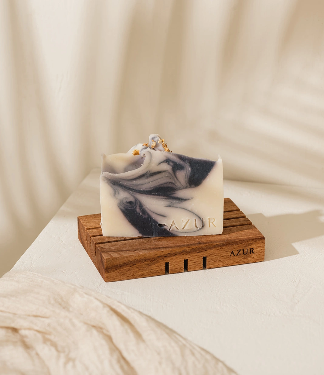 Wooden Soap tray from Azur Natural Body Care in use with their soap. Placed in a white setting, with shadows of a palm leaf in the background.