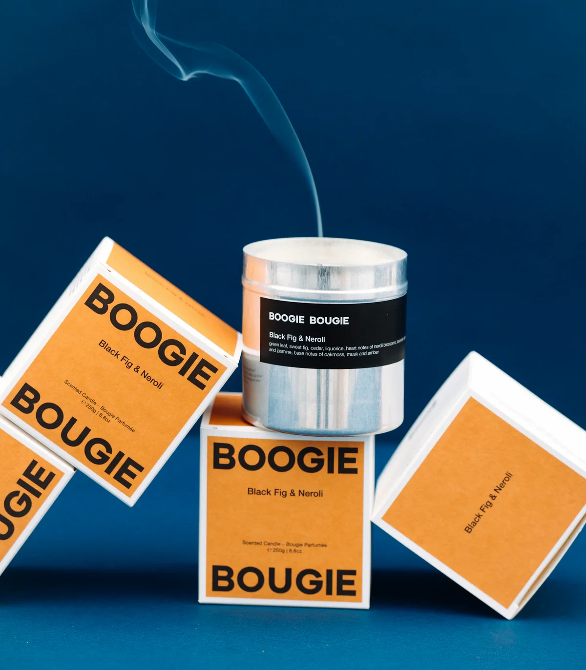 Boogie Bougie Black Fig & Neroli candle surrounded by its vibrant orange packaging. The aluminum candle container emits a thin wisp of smoke, adding a dynamic touch to the scene. Set against a deep blue background, the bold typography of the packaging creates a striking visual contrast.