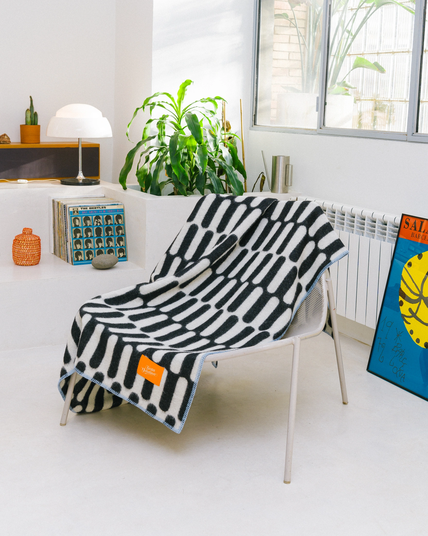 Throw Blanket From Tarta Gelatina coming in a black and white color. The pattern is similar to what you would see on op-art pictures. The edges are stiched with a light blue thread. In this picture the blanket is placed on a chair and is in a modern setting. 