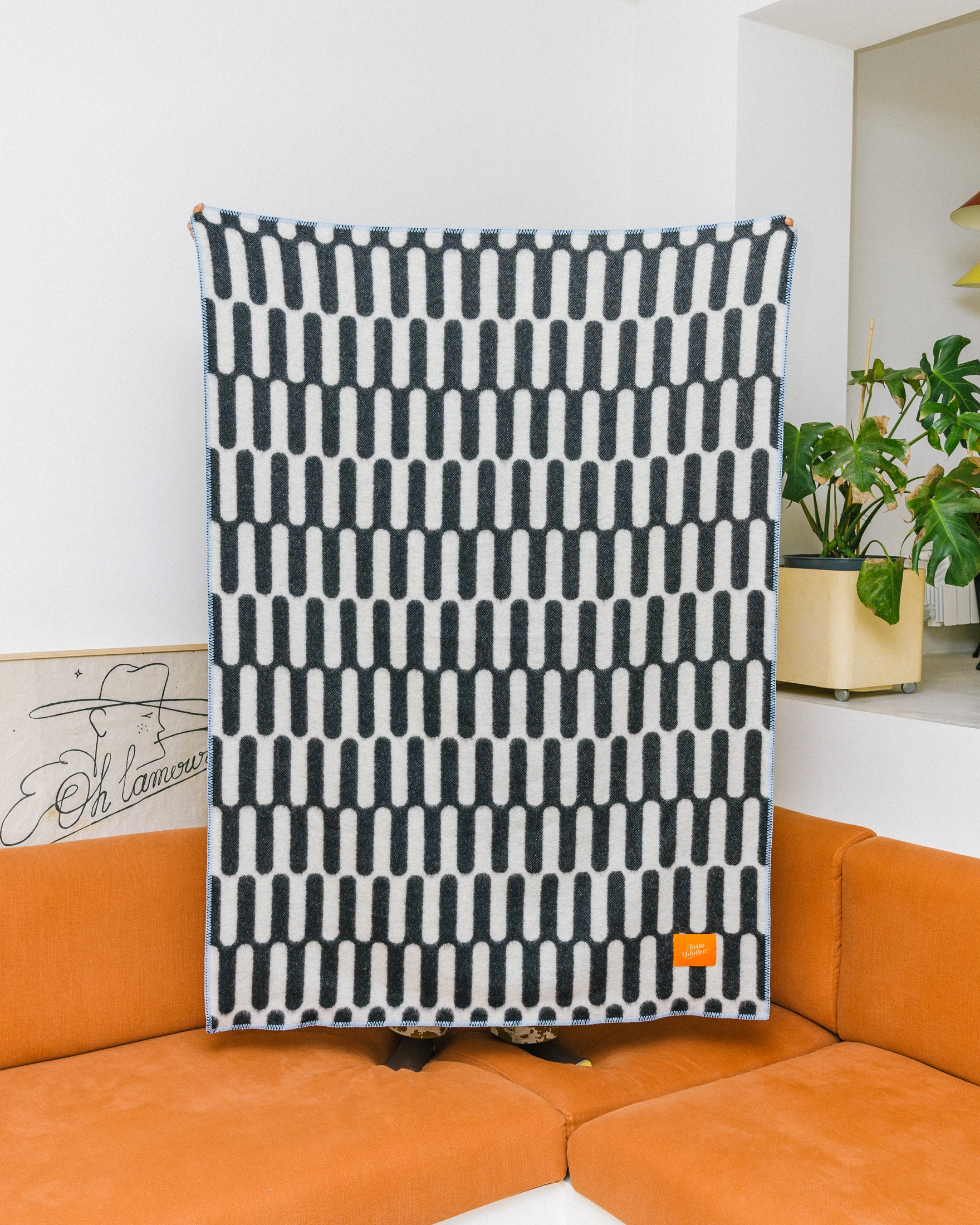 Throw Blanket From Tarta Gelatina coming in a black and white color. The pattern is similar to what you would see on op-art pictures. The edges are stiched with a light blue thread. In this picture the blanket held by someone so the whole pattern is visible.