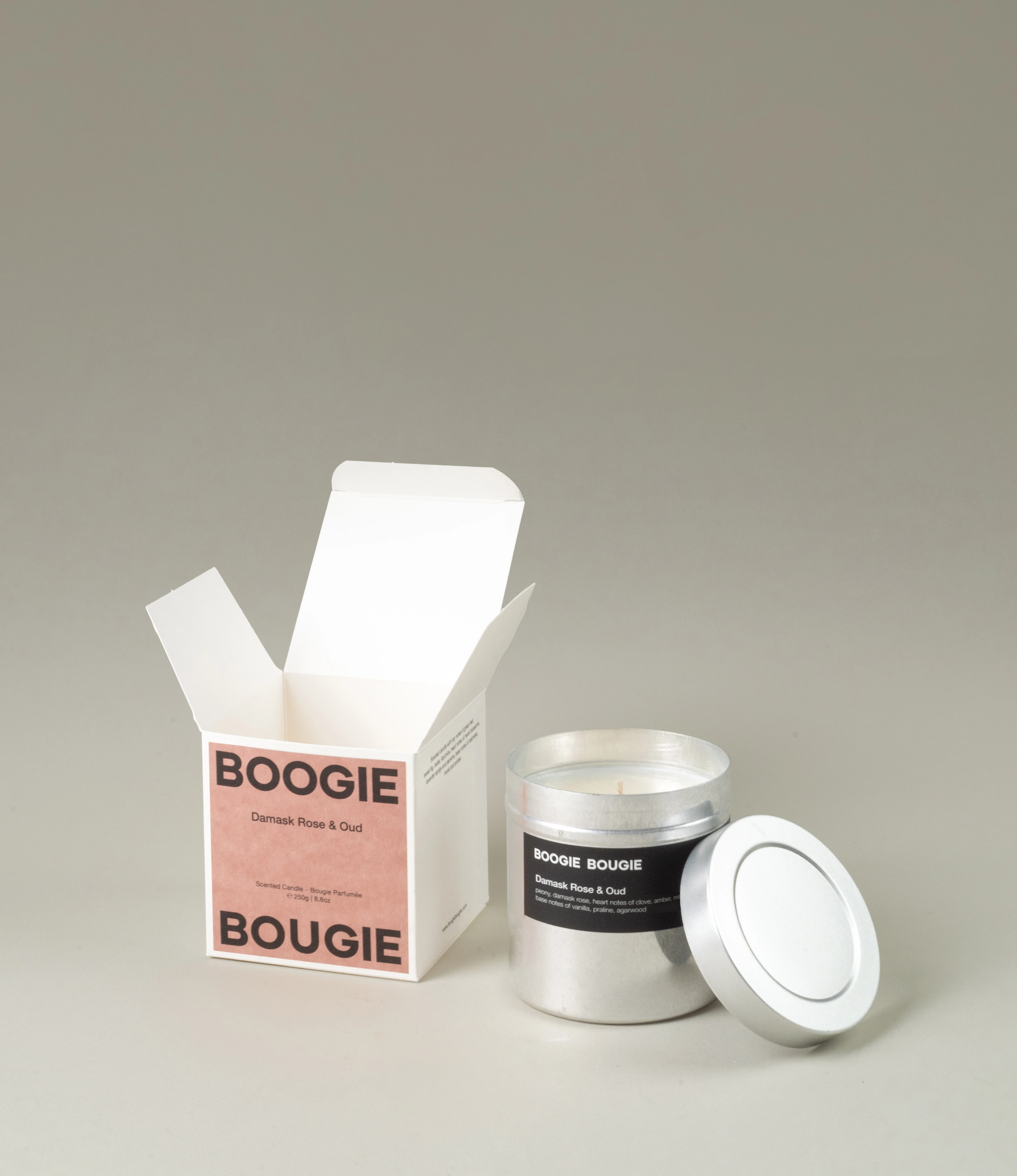 Boogie Bougie Damask Rose & Oud scented candle presented in a minimalistic aluminum container with a black label. The candle is placed next to its elegant pink and black branded packaging, with the box open to reveal the candle inside. A neutral background accentuates the sophisticated design.