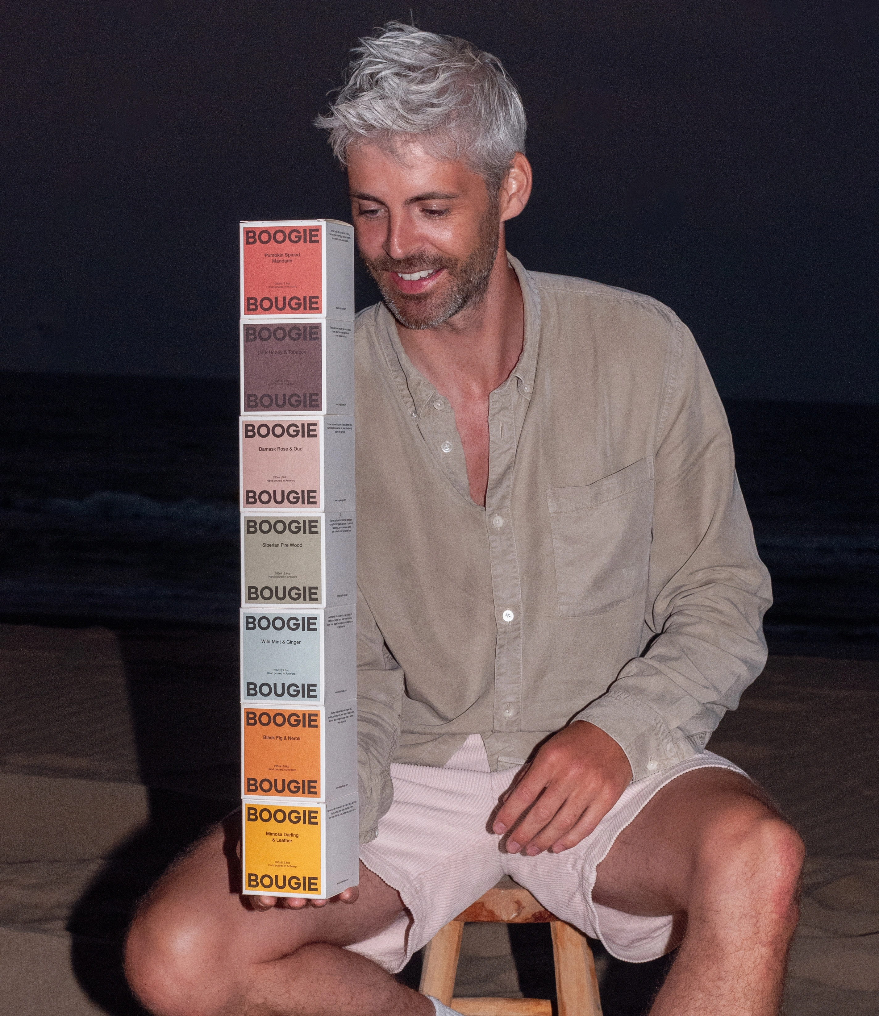 A man with silver hair, wearing a beige shirt and pink shorts, sitting on a wooden stool at the beach at night. He smiles while balancing a vertical stack of colorful Boogie Bougie candle boxes.