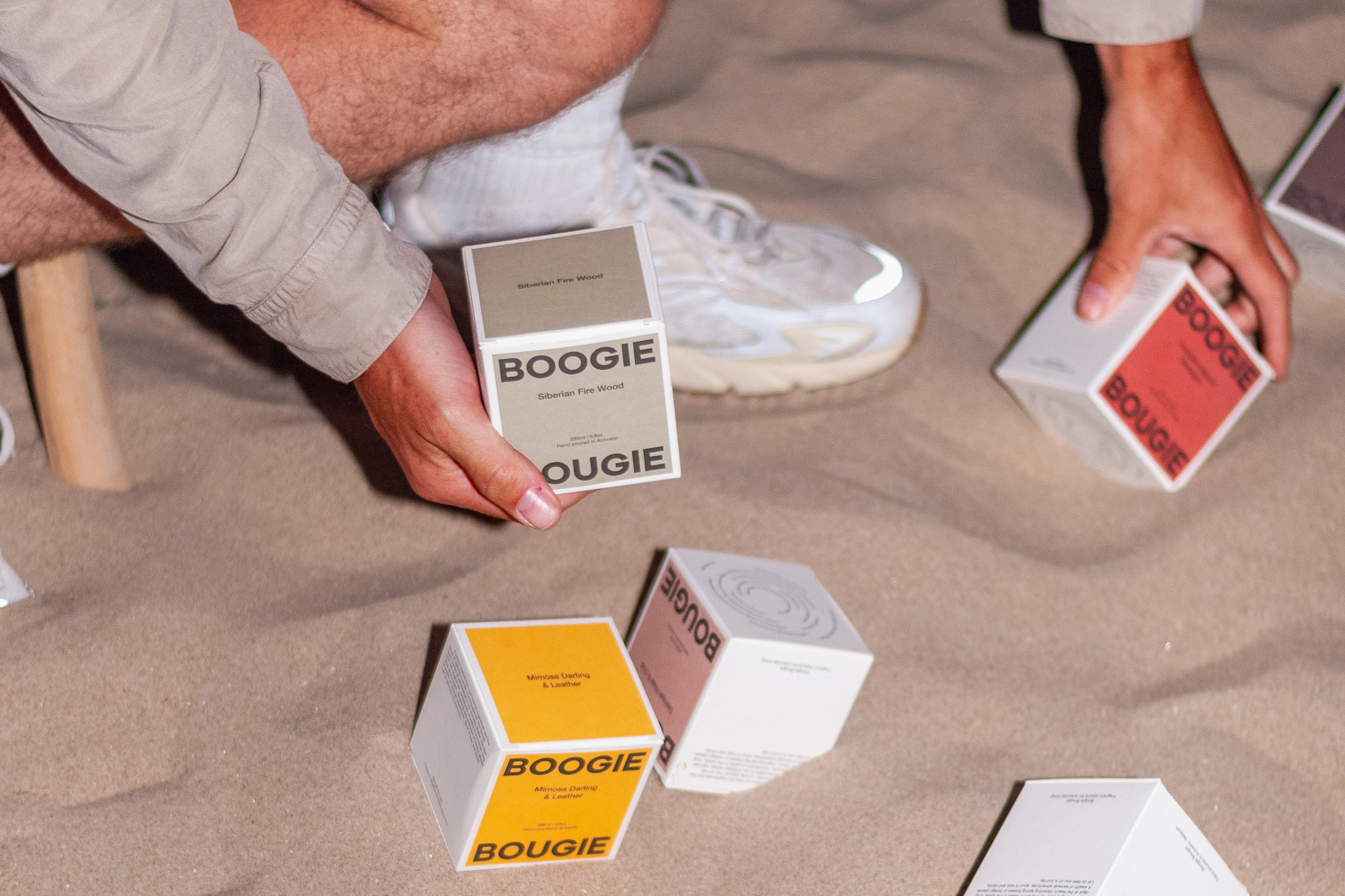 A person seated on a wooden stool on sand, holding a stack of colorful Boogie Bougie candle boxes, with additional candle boxes placed on the sand beside white sneakers and socks, shot during a nighttime beach setting.