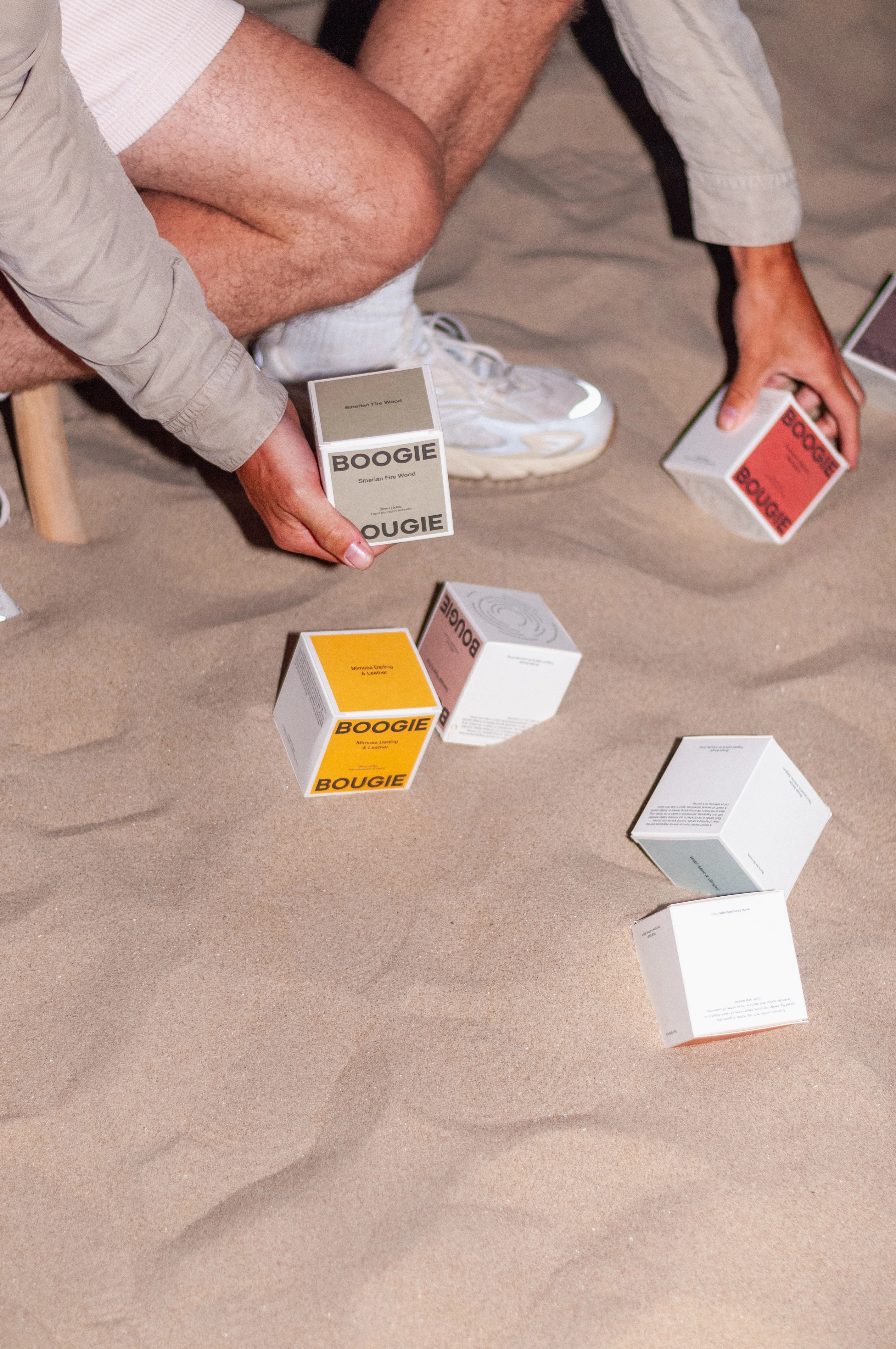 Person in a casual outfit kneeling in the sand, arranging BOOGIE candle boxes in various colors, with scattered boxes around.