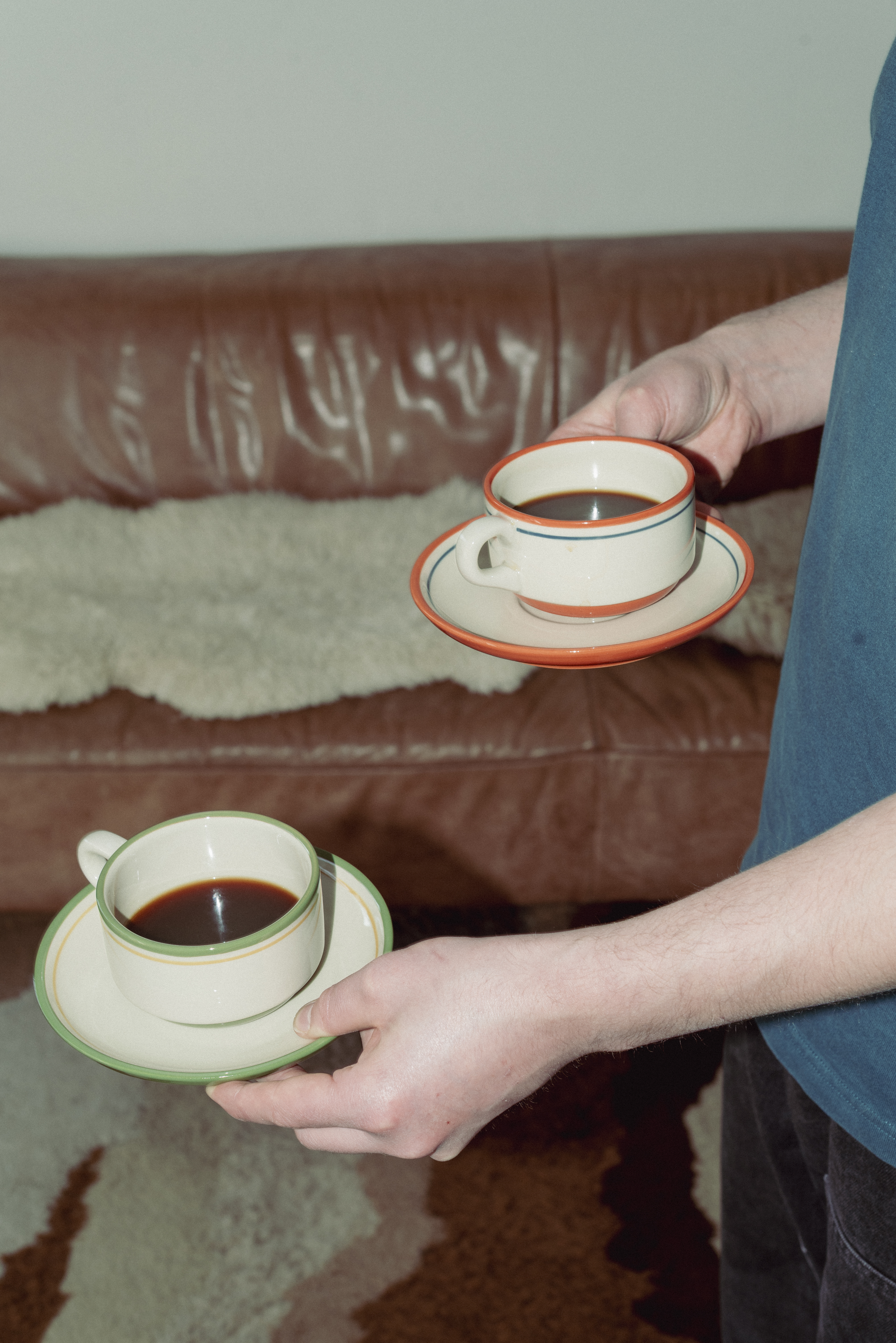 Person holding two Casa Atlântica stoneware coffee cups with saucers—one with orange and one with green hand-painted rims—filled with black coffee, in a warm interior setting with a leather sofa and wool rug in the background