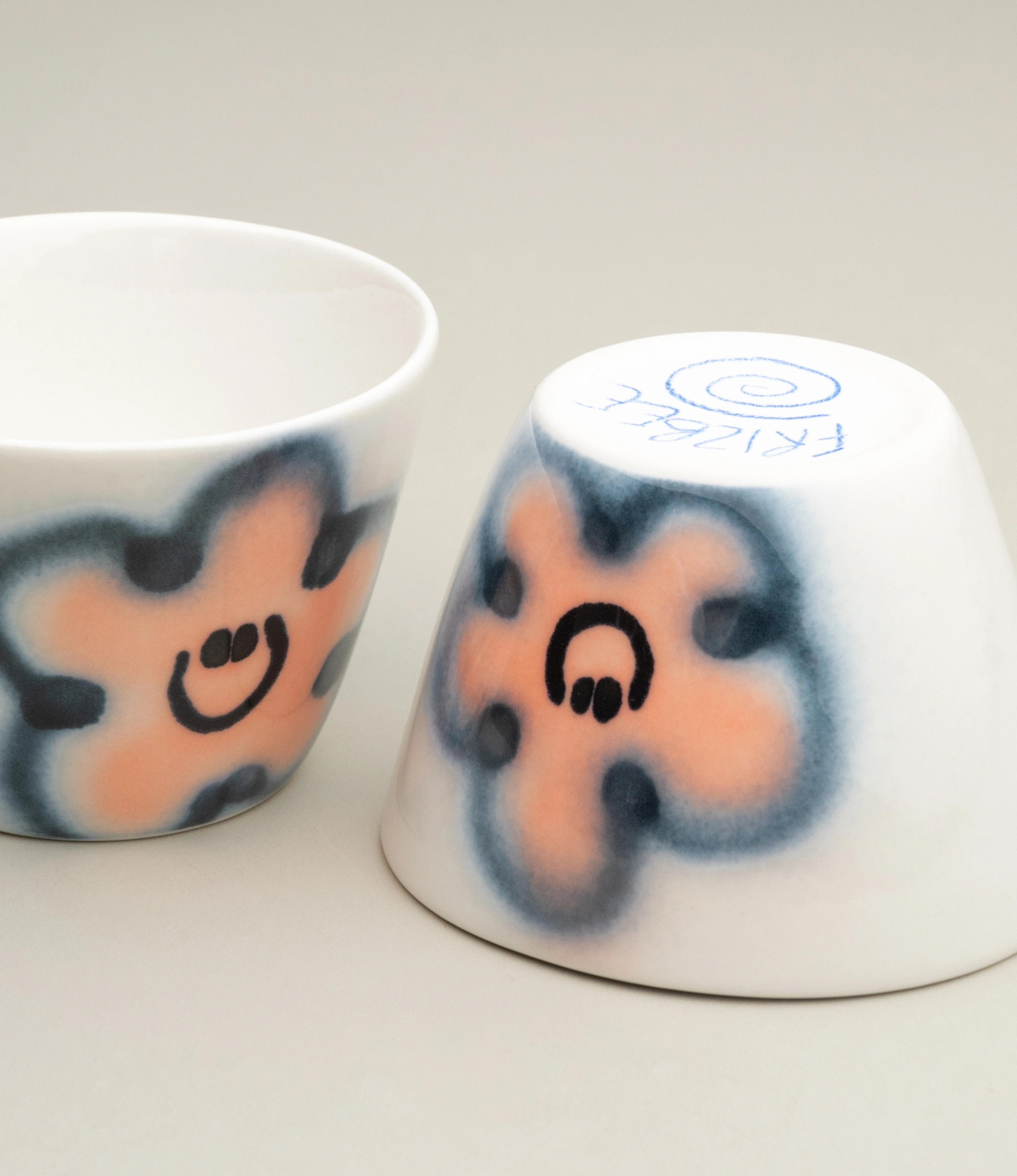 Close-up of the Frizbee Ceramics Super Cup in Peach Carne, highlighting the hand-painted smiley flowers in soft pink and dark blue hues. A cheerful and artistic ceramic piece.