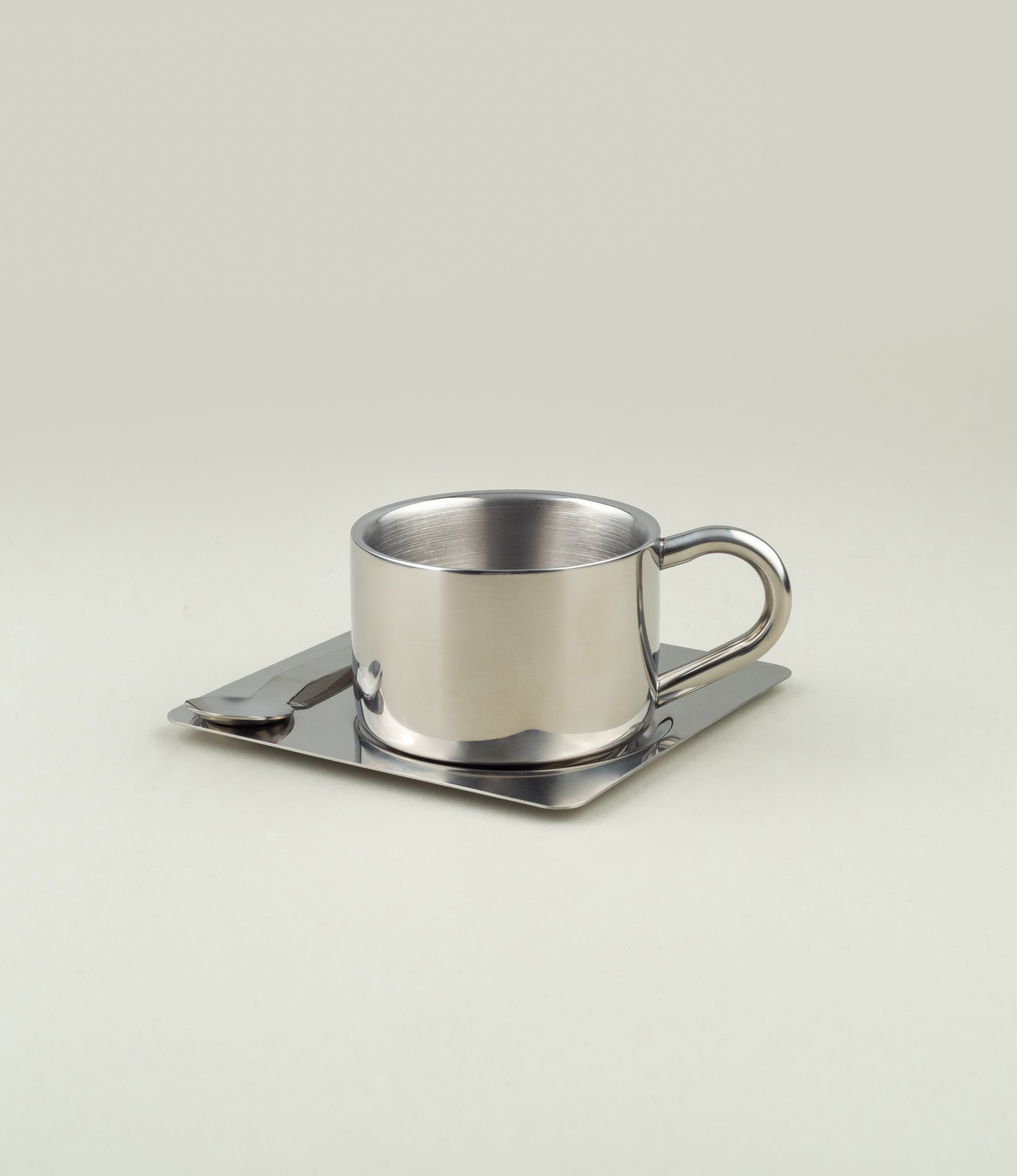 A stainless steel cup with a U-shaped handle sits on a square stainless tray; a flat-handled spoon rests beside the cup, all on a pale background.
