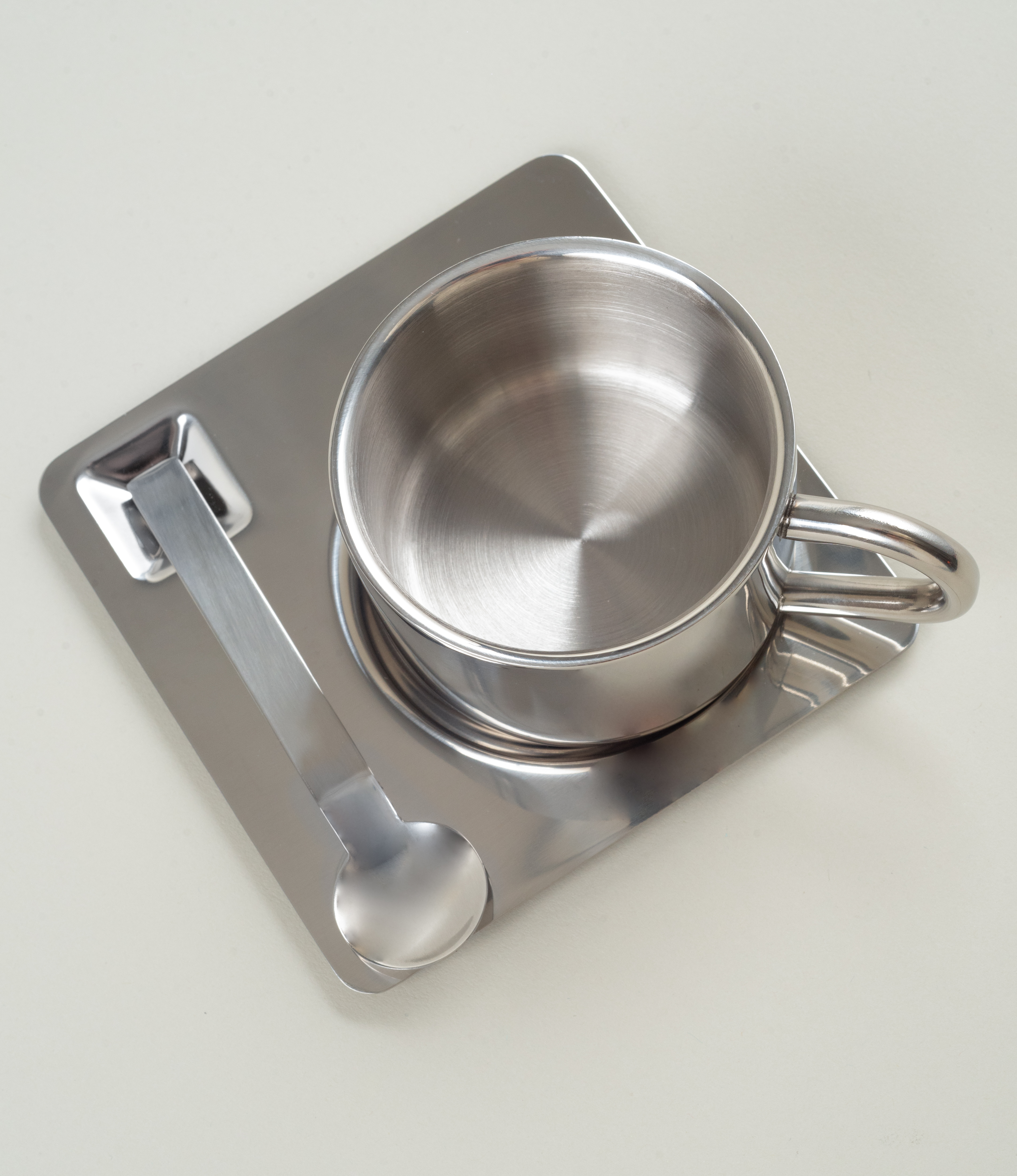 Top-down shot focused on the concentric brush marks inside the stainless steel cup, the tray beneath it and the spoon positioned to the left.