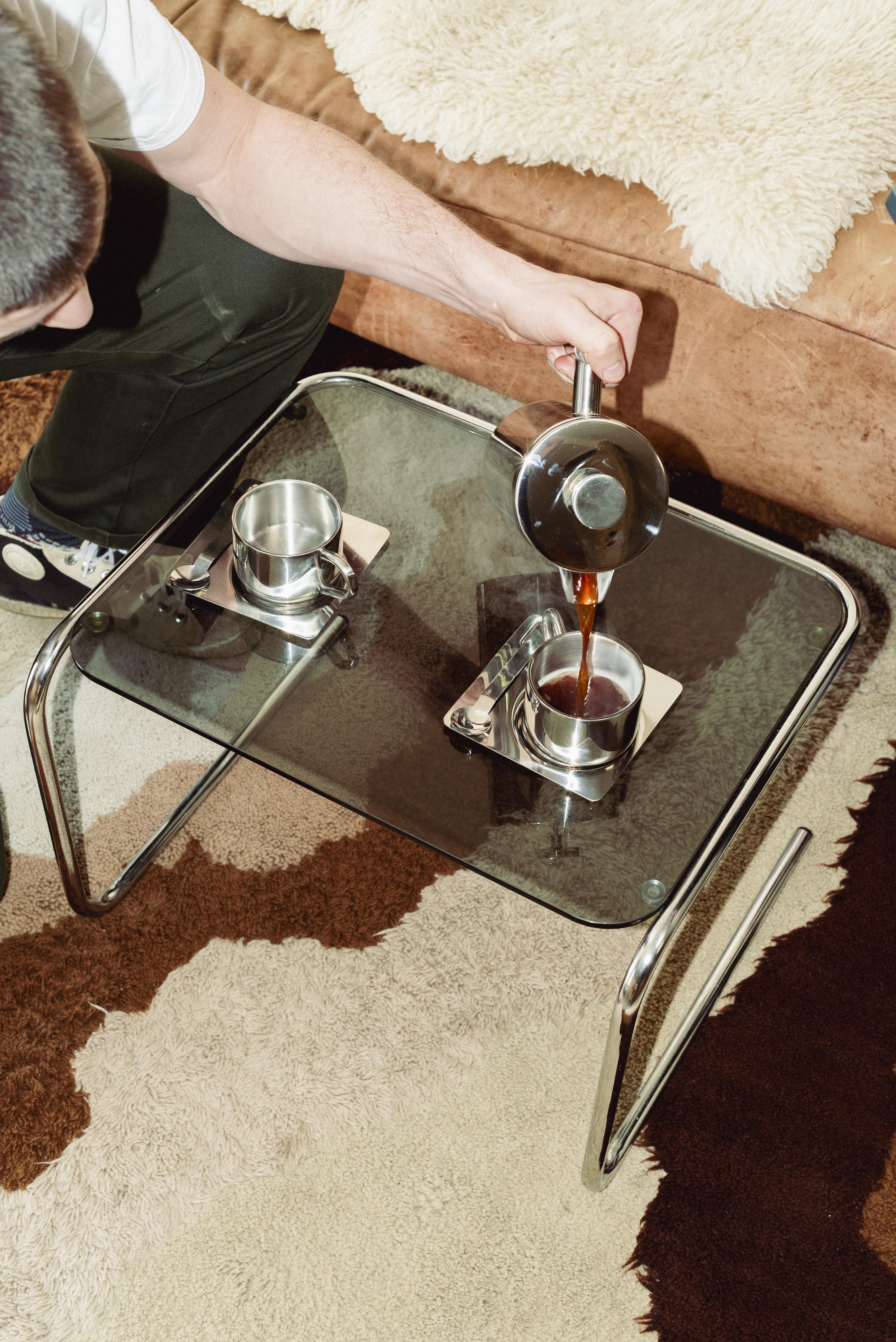 Overhead perspective of a hand pouring coffee from a stainless steel French press into one of two stainless steel cups on square trays with spoons, on a glass-top table in front of a leather couch and sheepskin throw.