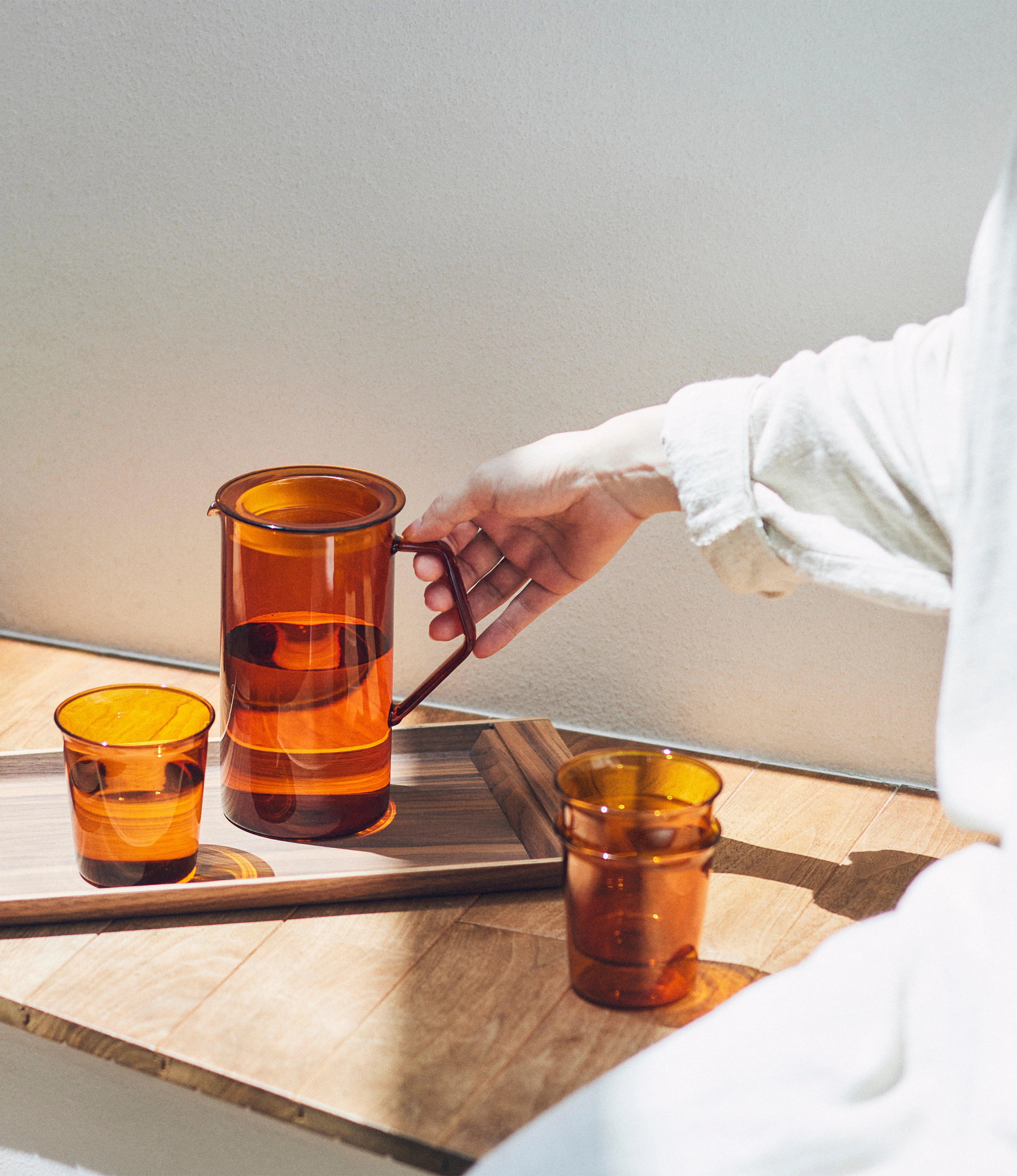 Cast Amber Jug and Glass from Kinto. The Item can hold 750ml of liquid. The modern geometric shape is a perfect companion to any kitchen. In this picture the products are in use, the jug is held by a person dressed in white linen clothes.