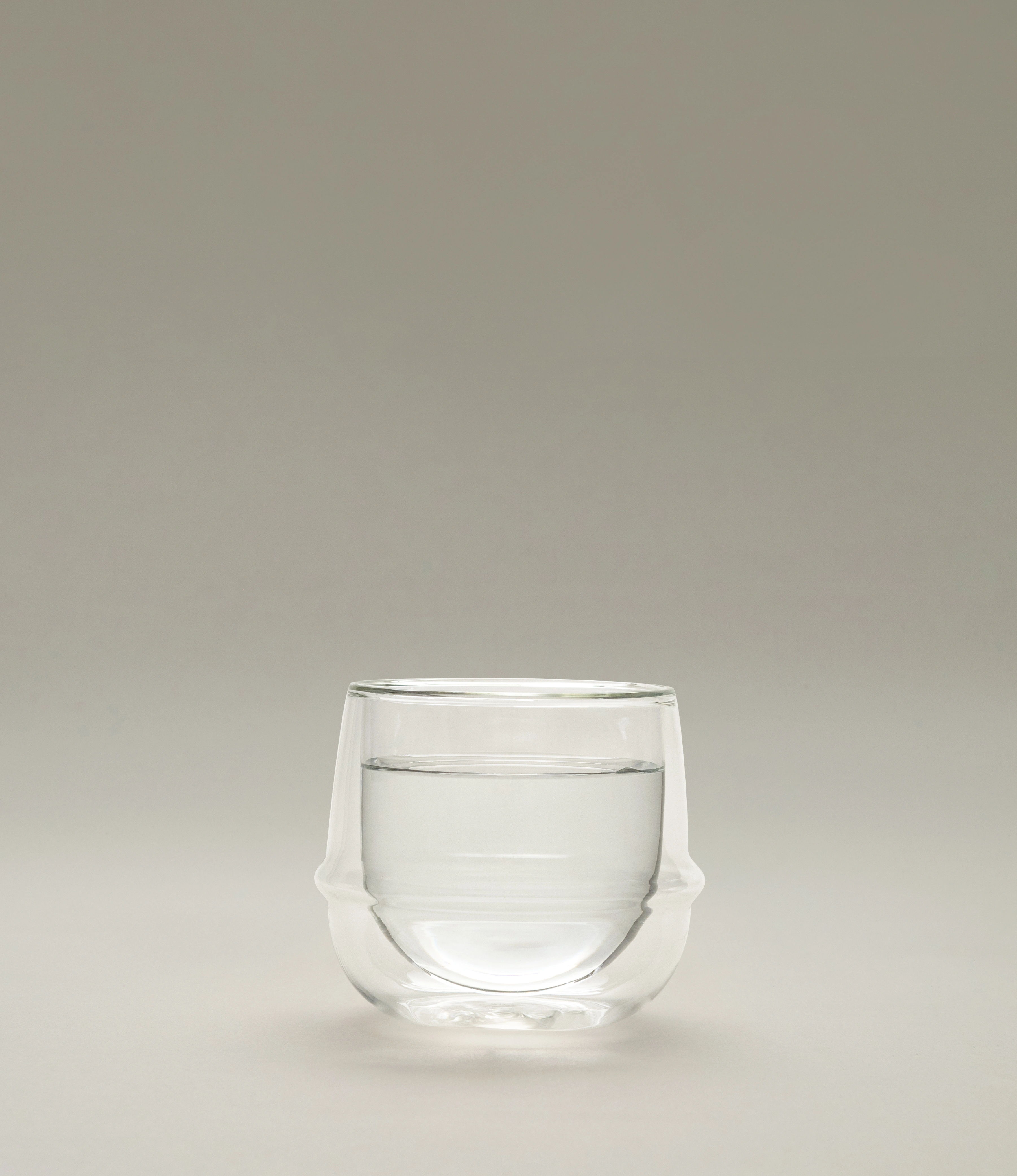 A minimalist double-walled glass filled with water, sitting on a neutral background. The glass has a unique curvilinear design with an insulating air layer, highlighting its elegant and functional form. The soft light enhances its clarity and craftsmanship.