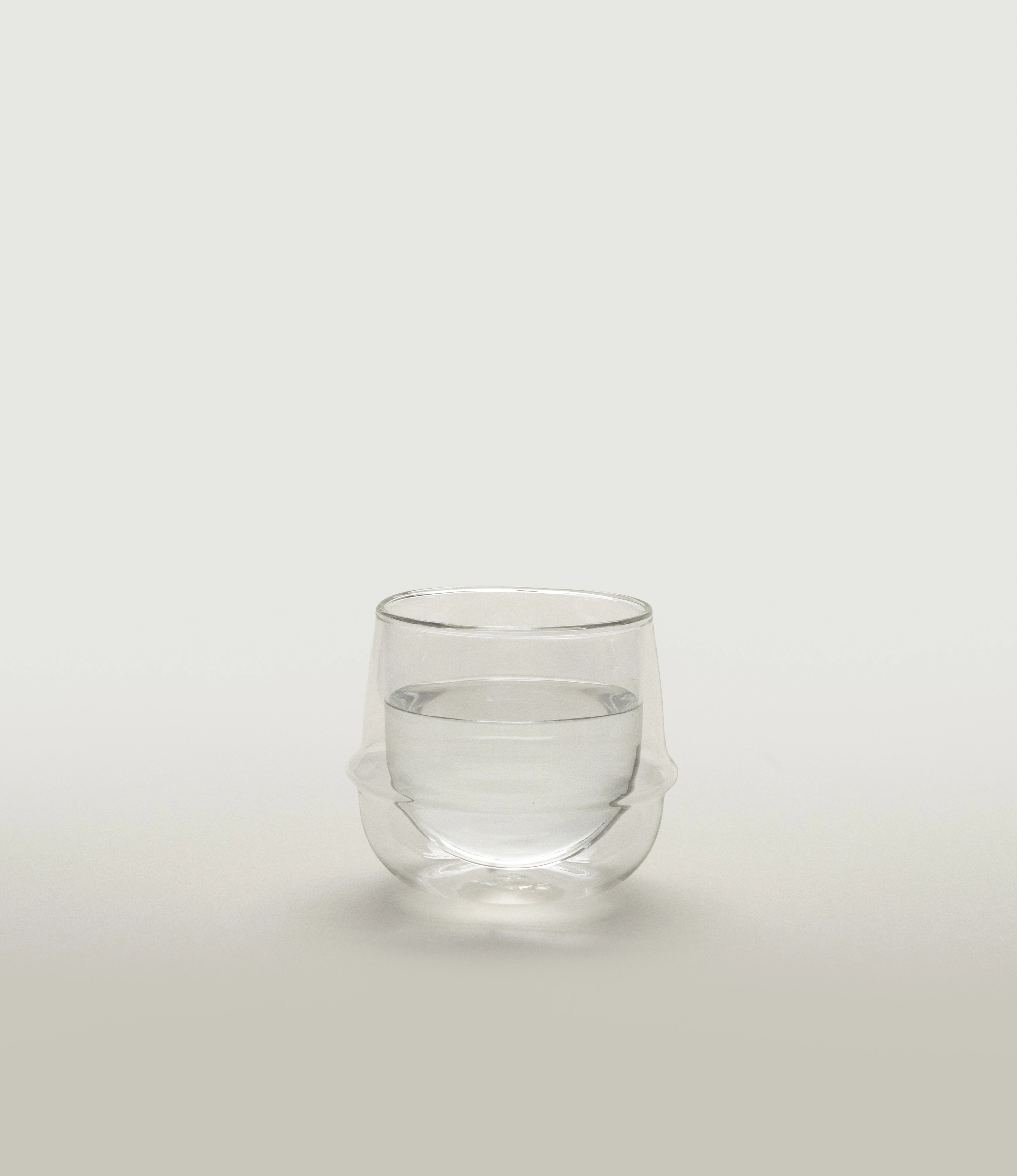 A minimalist double-walled glass filled with water, sitting on a neutral background. The glass has a unique curvilinear design with an insulating air layer, highlighting its elegant and functional form. The soft light enhances its clarity and craftsmanship.