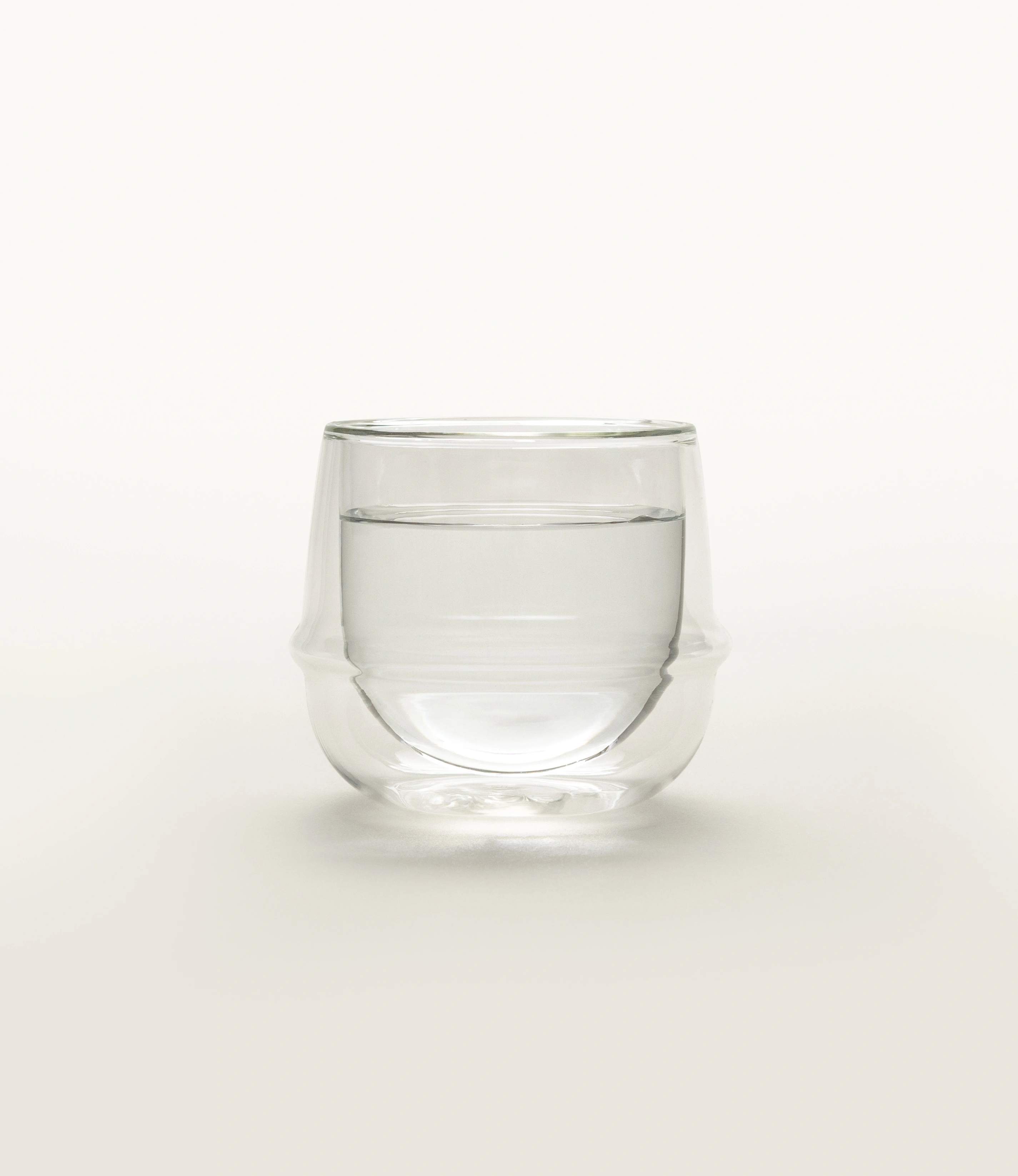 A minimalist double-walled glass filled with water, sitting on a neutral background. The glass has a unique curvilinear design with an insulating air layer, highlighting its elegant and functional form. The soft light enhances its clarity and craftsmanship.