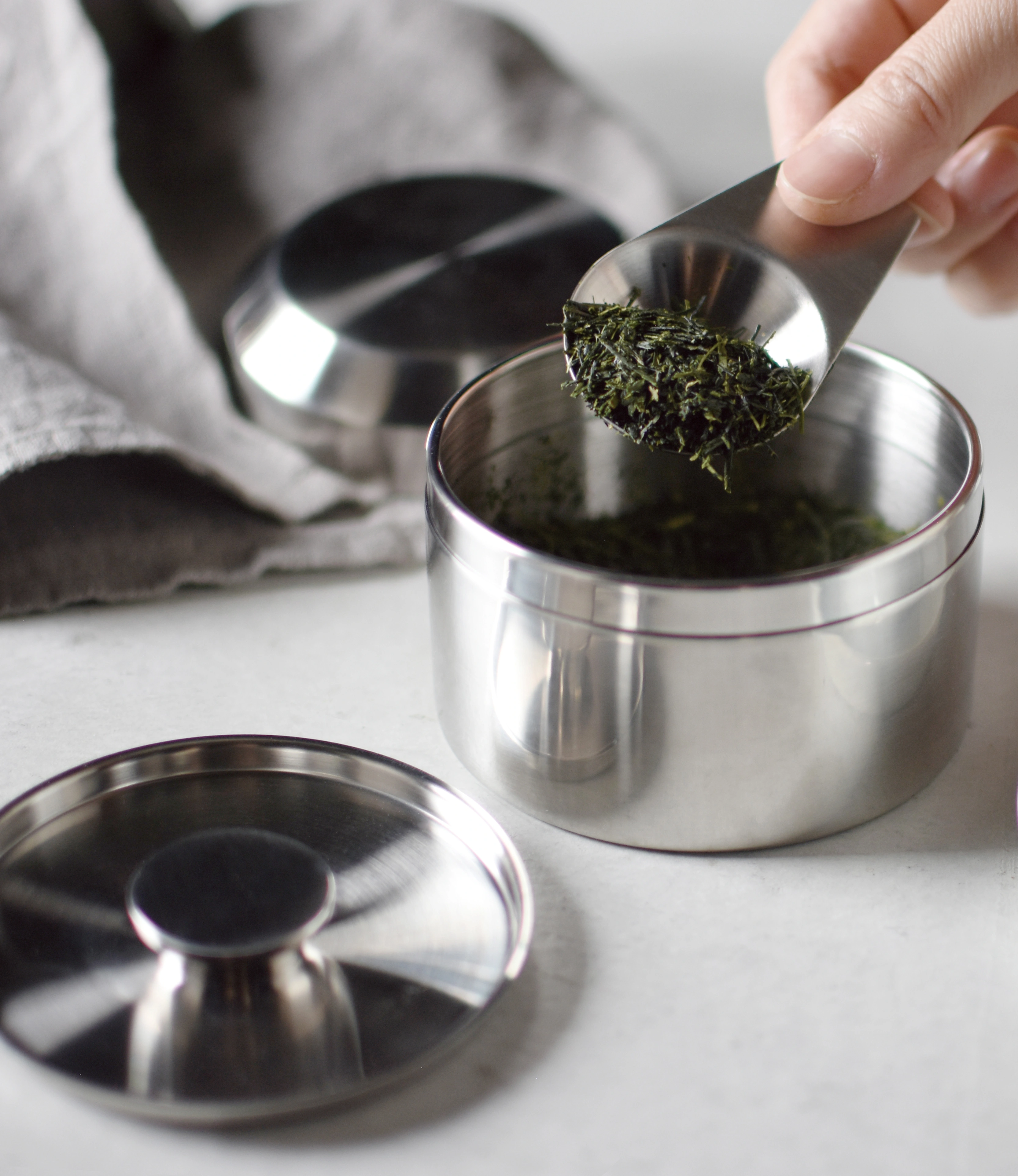 LT Tea Scoop in use with LT Tea Canister S. Both of the Items are from the japanese brand Kinto and are made of stainless steel. The item is being held by a hand.