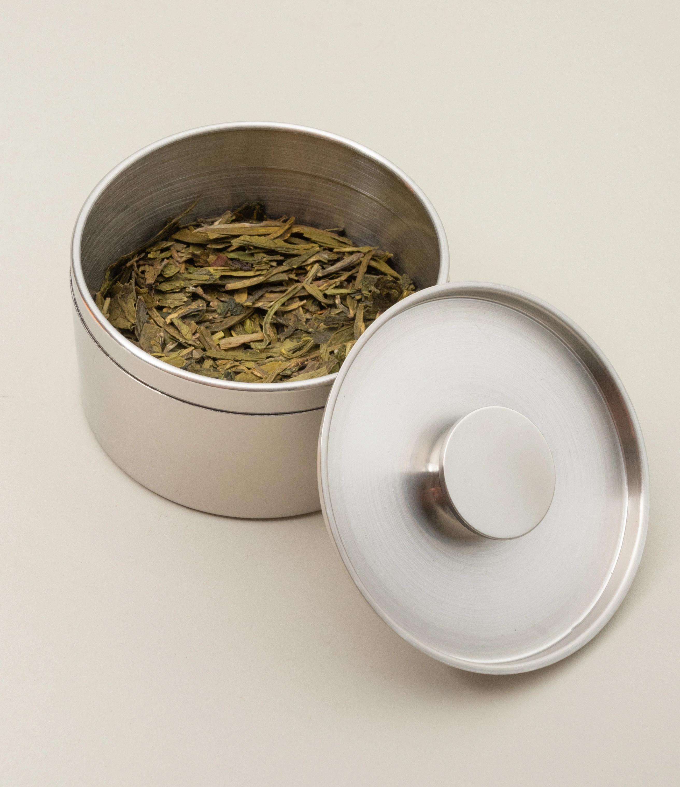 Close-up of an open stainless steel tea canister filled with loose-leaf green tea. The canister's brushed metal inner surface reflects light softly, while the lid with a central knob handle is placed on its side. The neutral background emphasizes the elegant and functional design.