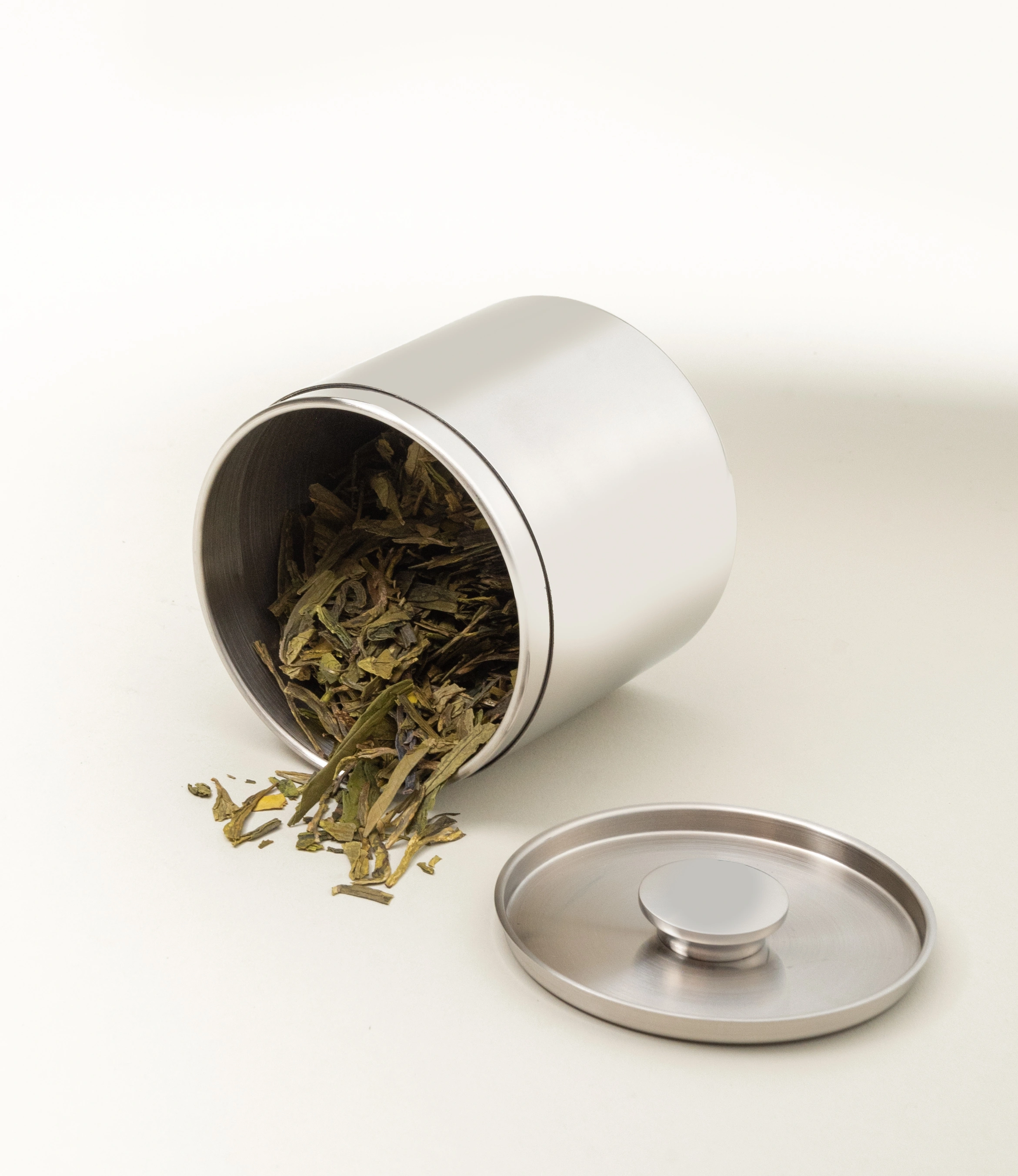 A stainless steel tea canister tipped on its side with loose-leaf green tea spilling out onto a neutral background. The detached lid with a circular knob handle lies flat in the foreground, showcasing its brushed metal finish. The minimalist design highlights functionality and aesthetic appeal.