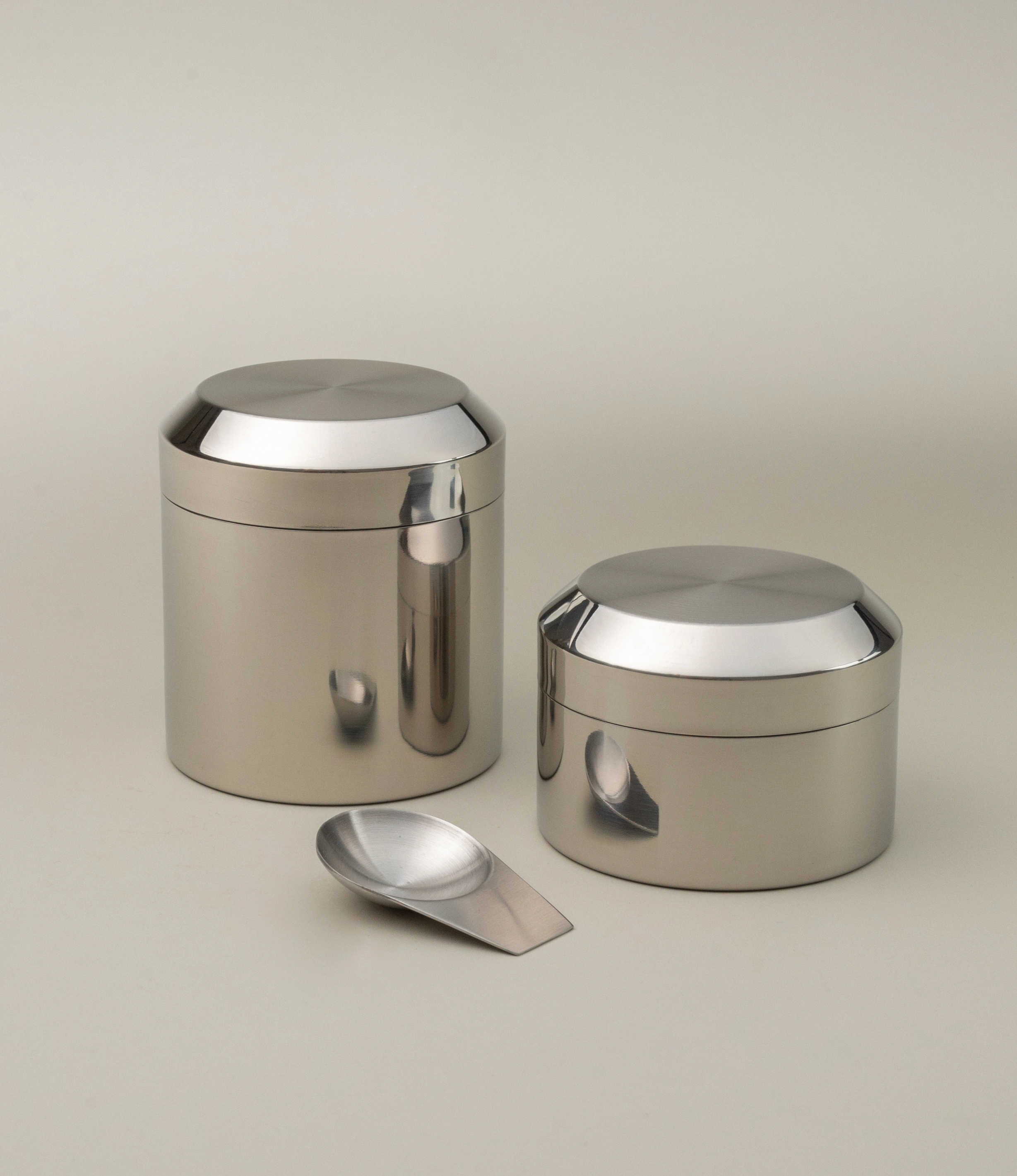Sleek stainless steel tea canister set with a polished finish, featuring one tall and one shorter container. Both have rounded lids that seamlessly integrate with the cylindrical bodies, reflecting a minimalist design. A matching metallic scoop is placed in front, completing the set. The ensemble is set against a neutral background to emphasize its modern and elegant aesthetic.