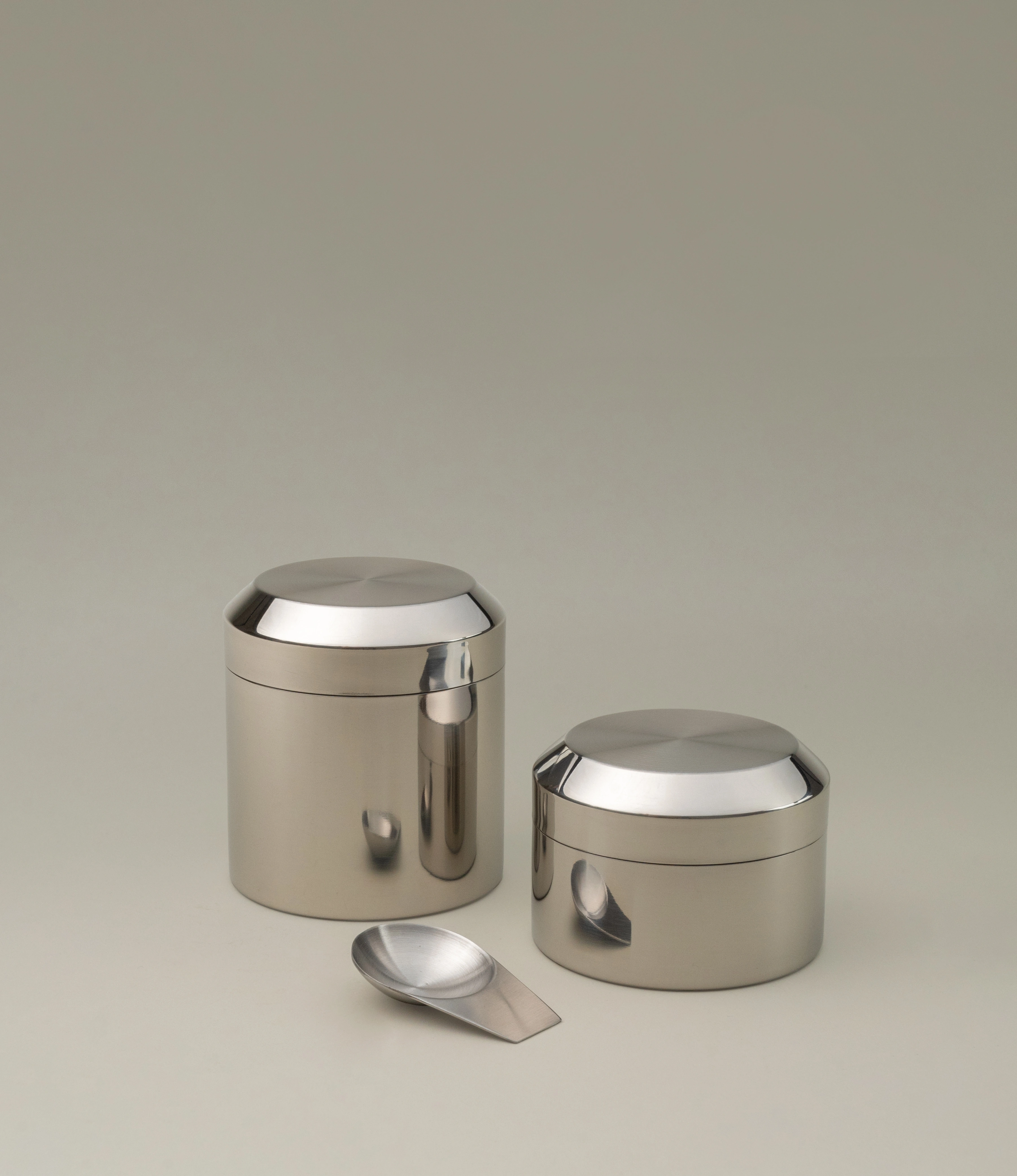 Sleek stainless steel tea canister set with a polished finish, featuring one tall and one shorter container. Both have rounded lids that seamlessly integrate with the cylindrical bodies, reflecting a minimalist design. A matching metallic scoop is placed in front, completing the set. The ensemble is set against a neutral background to emphasize its modern and elegant aesthetic.