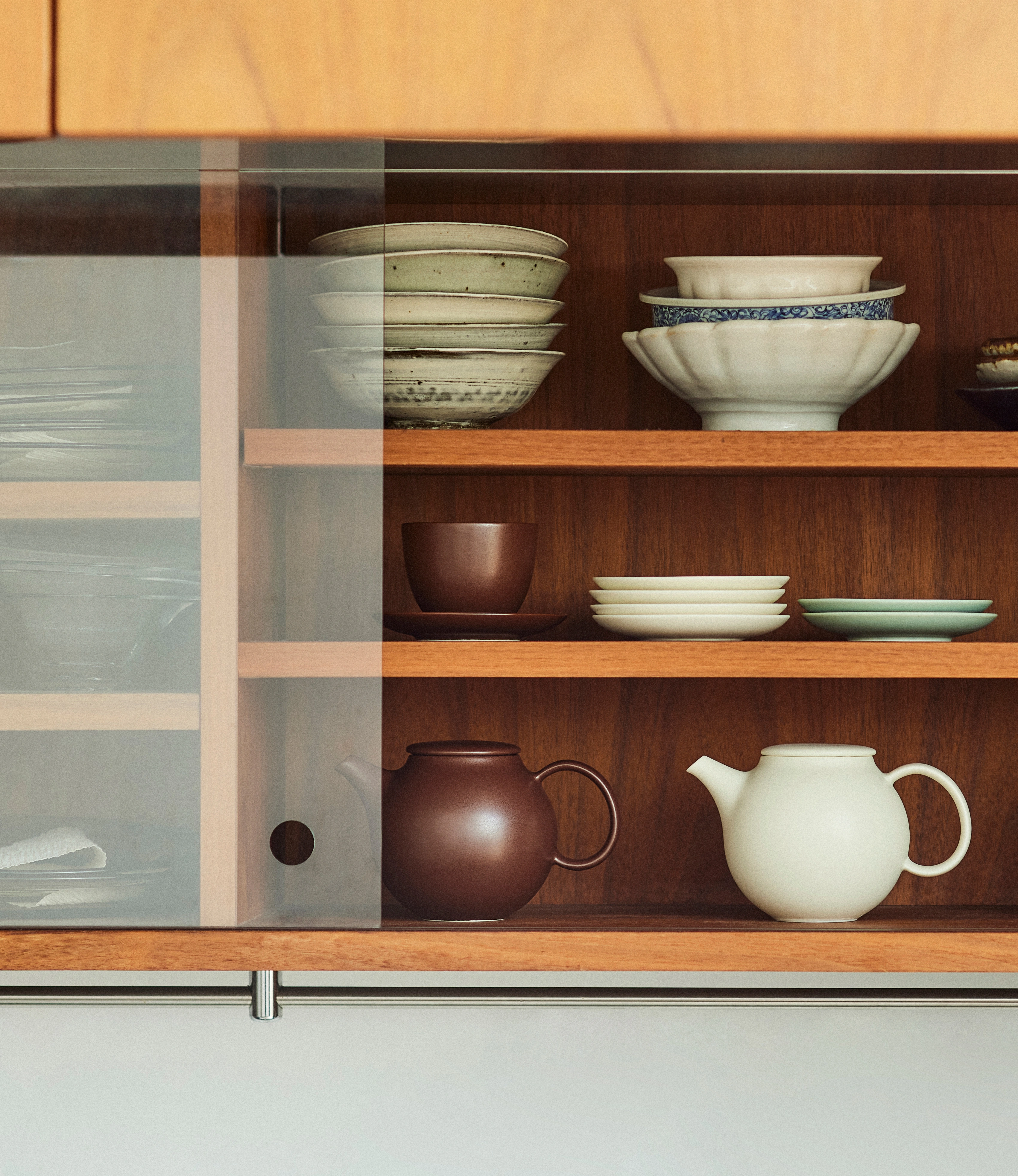 Pebble Tea Pot Brown in situation picture. The product is placed on a cupboard with a glass front with some other items like the white version of the exact same teapot.