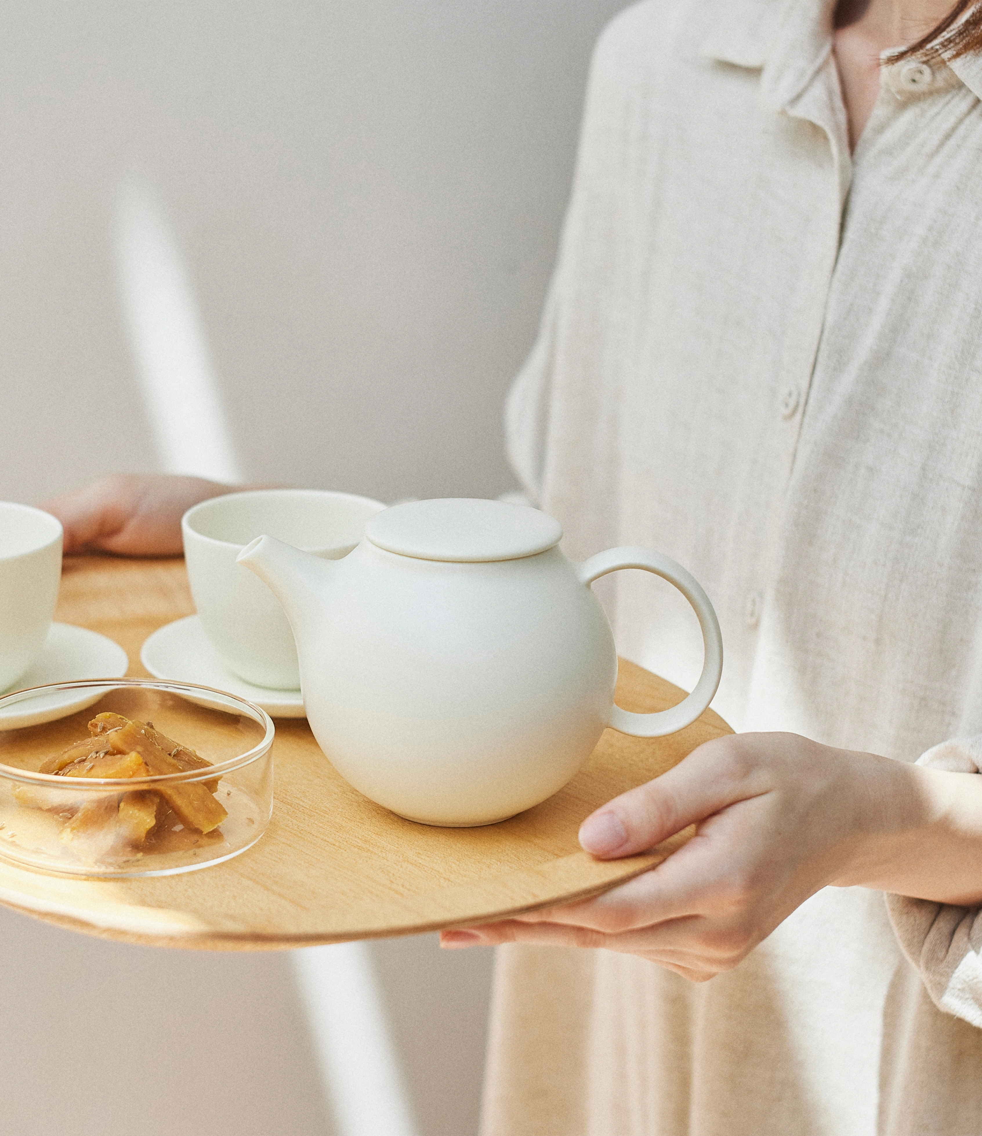 Pebble Tea Pot from Kinto in use. The item is placed on a wooden tray and it is held by a person dressed in beige linen clothes. There are some biscuits and cups next to it.