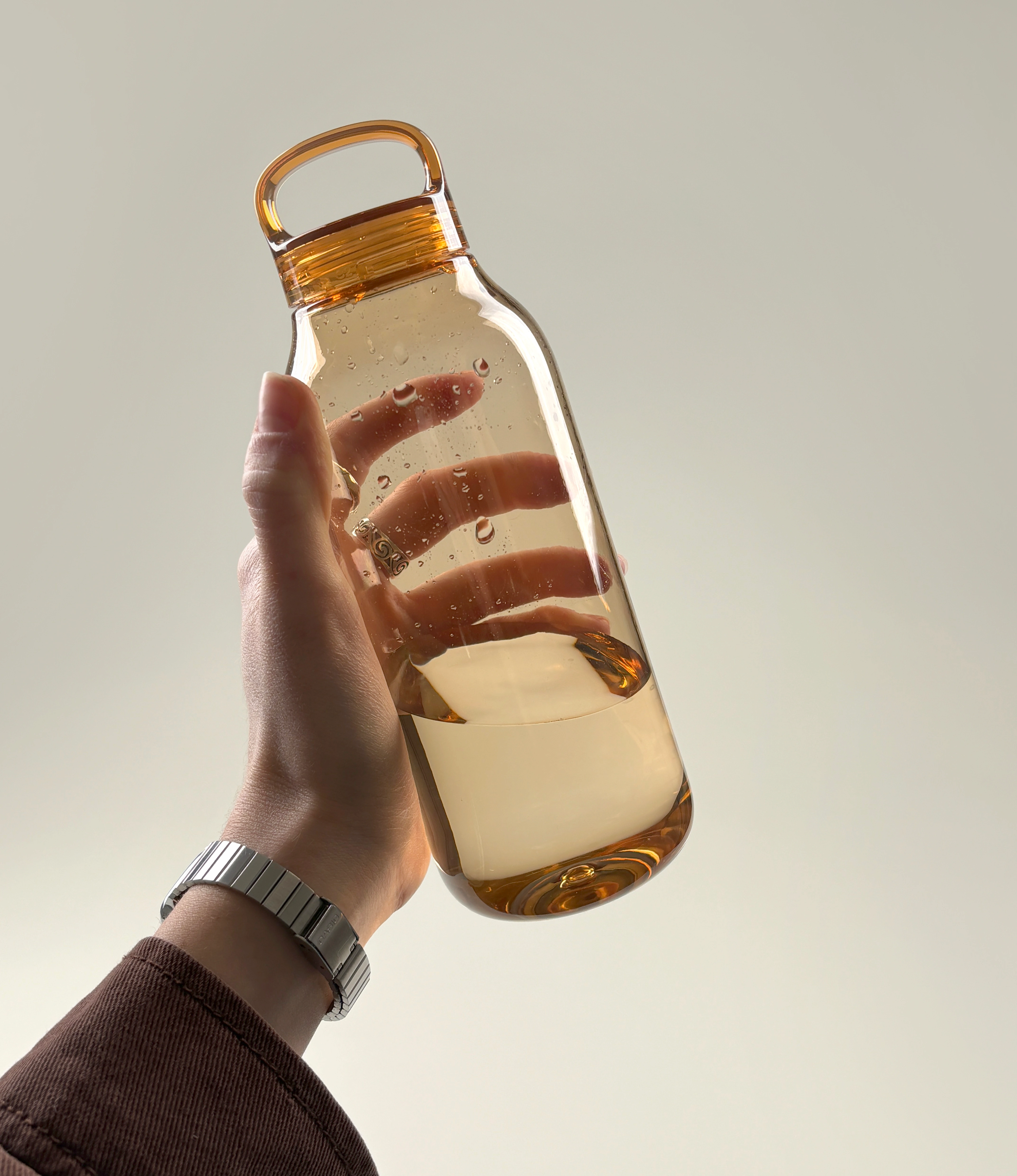 Hand holding an amber-colored water bottle partially filled with water, showcasing the sleek design and ergonomic handle. Droplets of water are visible on the bottle's exterior, adding a dynamic and refreshing feel.