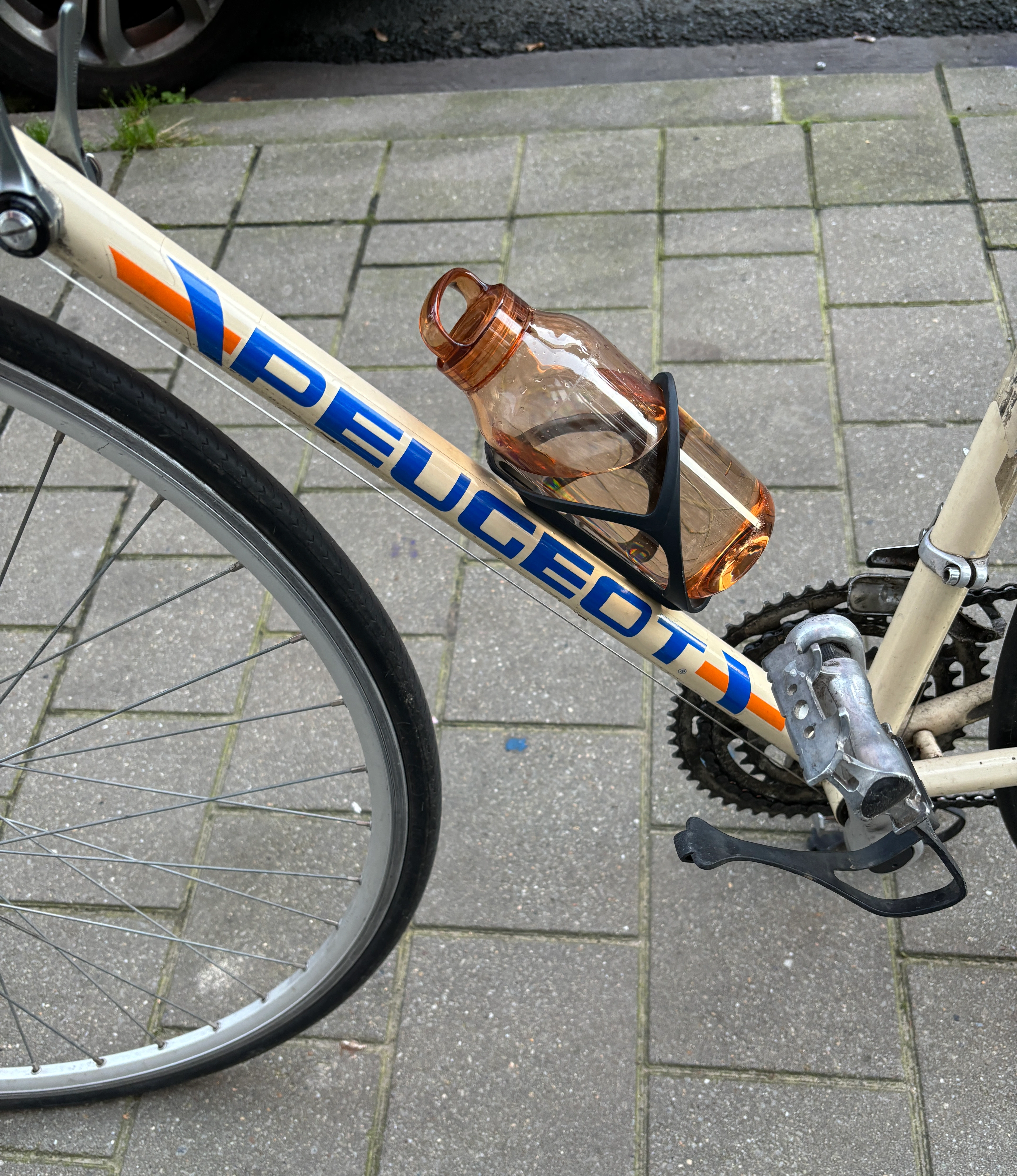 A vintage Peugeot bicycle with an amber-colored water bottle secured in a bottle holder on the bike frame, positioned on a tiled pavement.