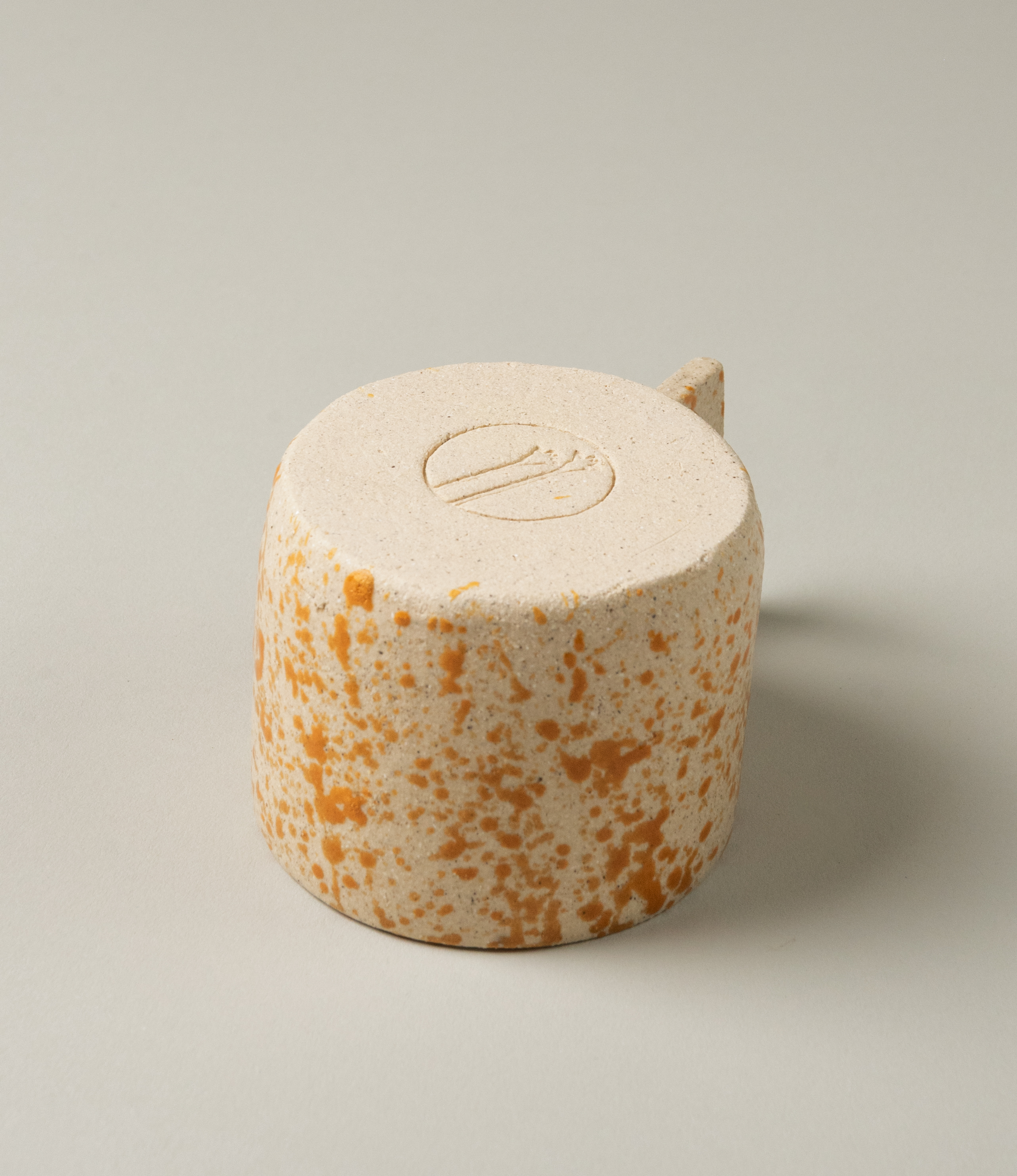 Pumpkin Spice Coffee Cup from Manamo Cups. The product has a natural touch and a modern shape with the rectangular handle. The item has a spotted orange texture on a beige base. The bottom of the product wears the debossed logo of Manamo Cups. 