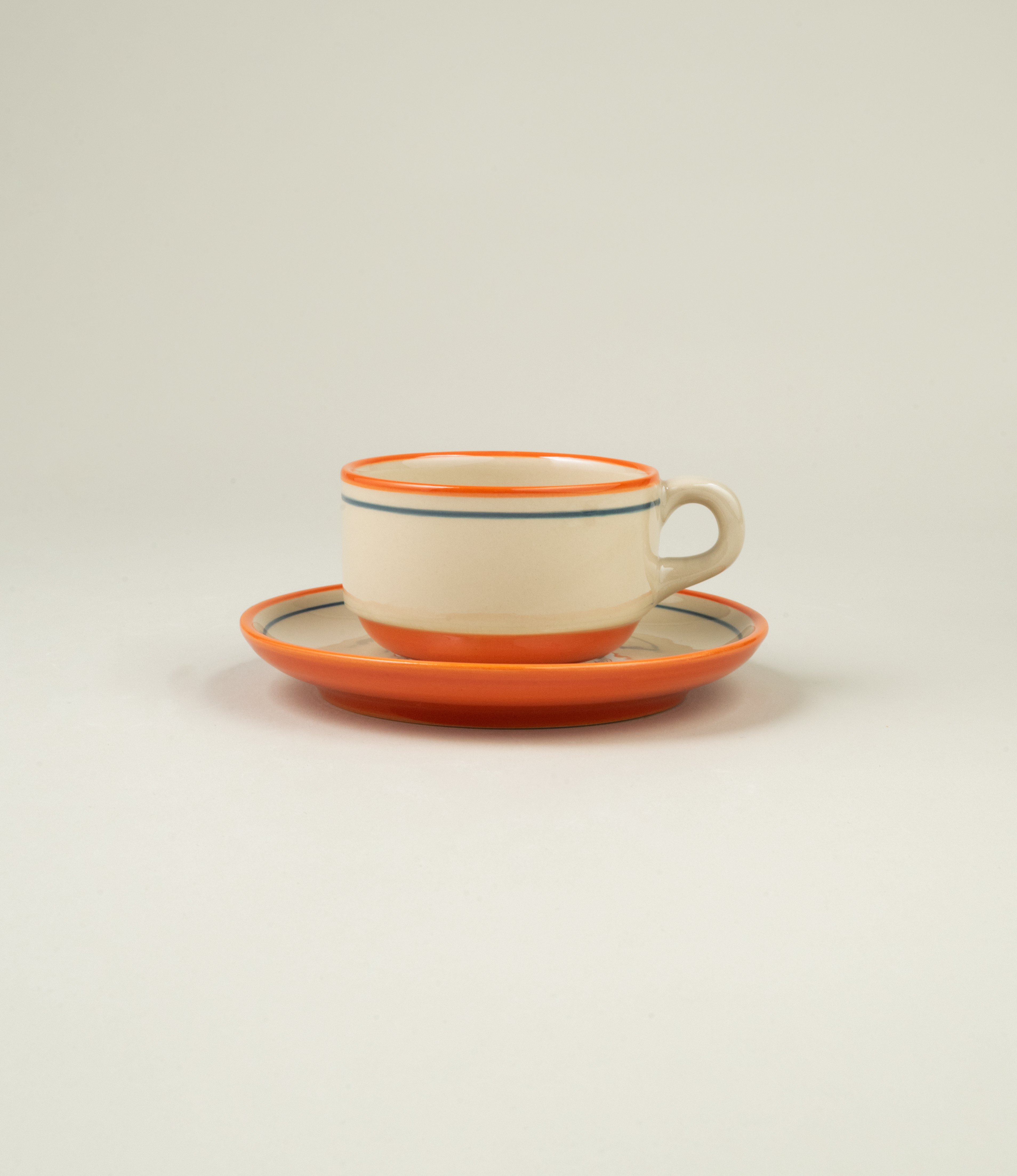 A frontal view of a stoneware cup and saucer with hand-painted orange and blue lines, inspired by Spanish café culture.