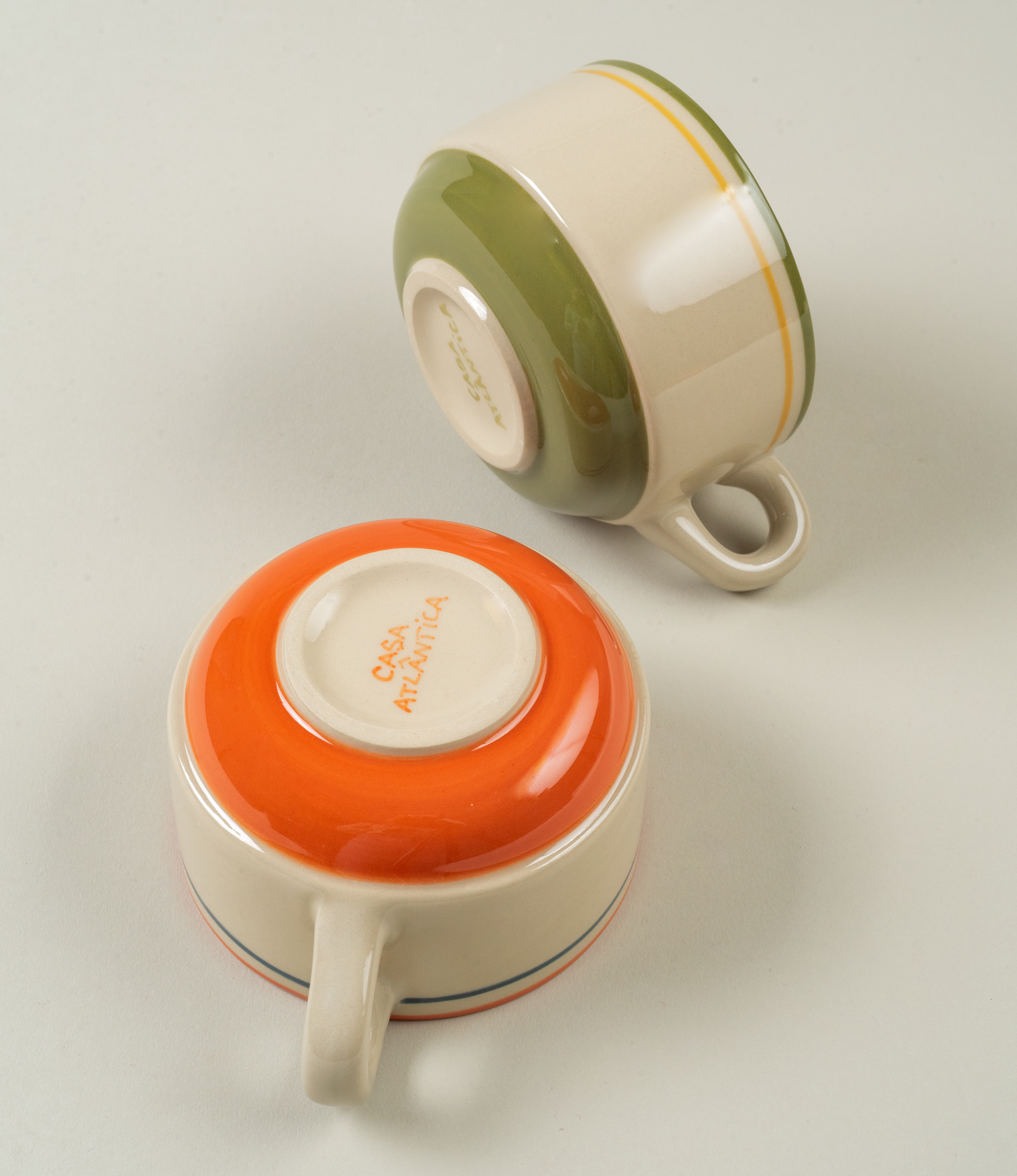 Two Casa Atlântica stoneware coffee cups, one with orange and the other with green accents, showcasing hand-painted glazes.