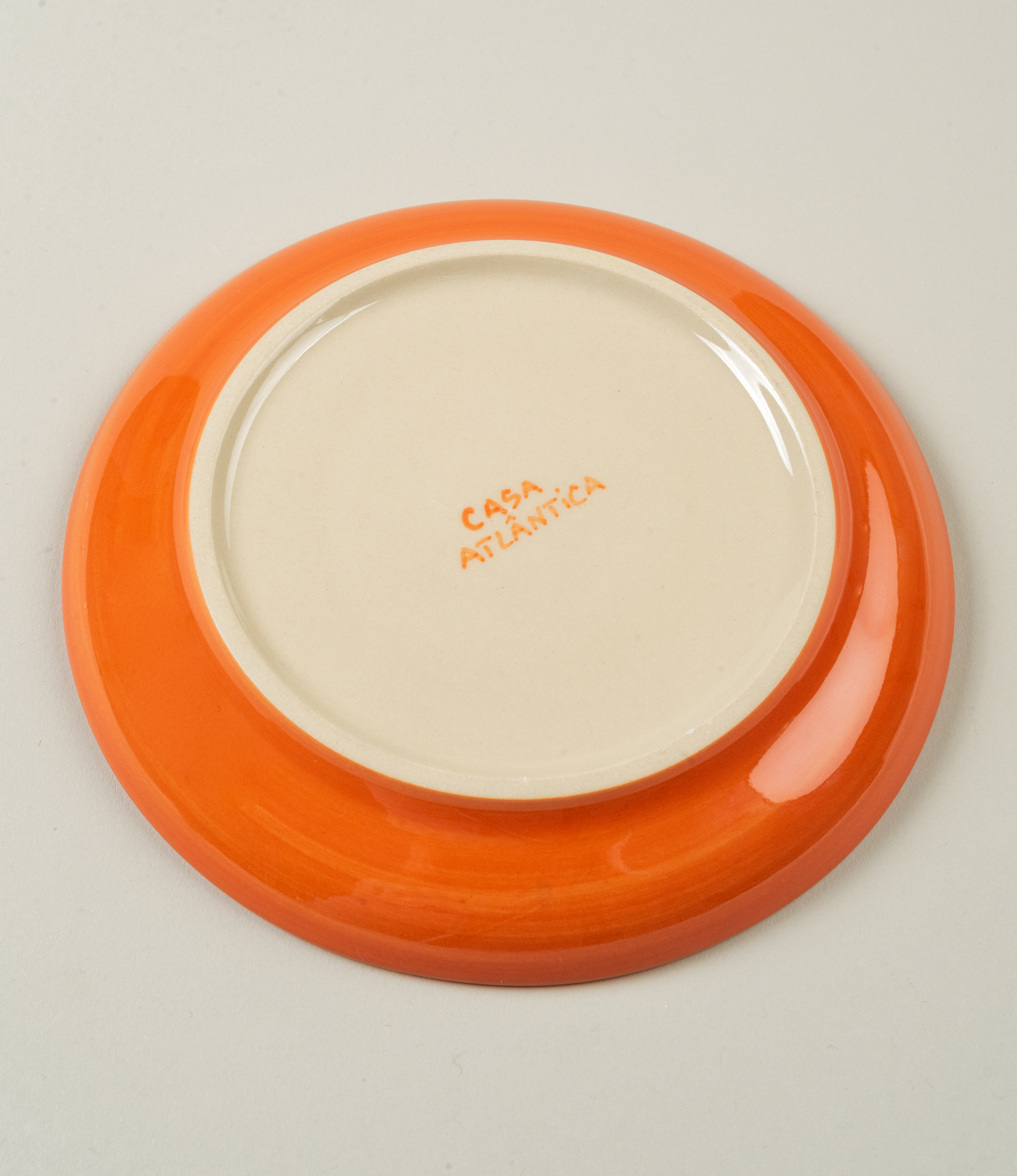 Bottom view of Casa Atlântica saucer with orange glazed rim and hand-painted brand name in the center.