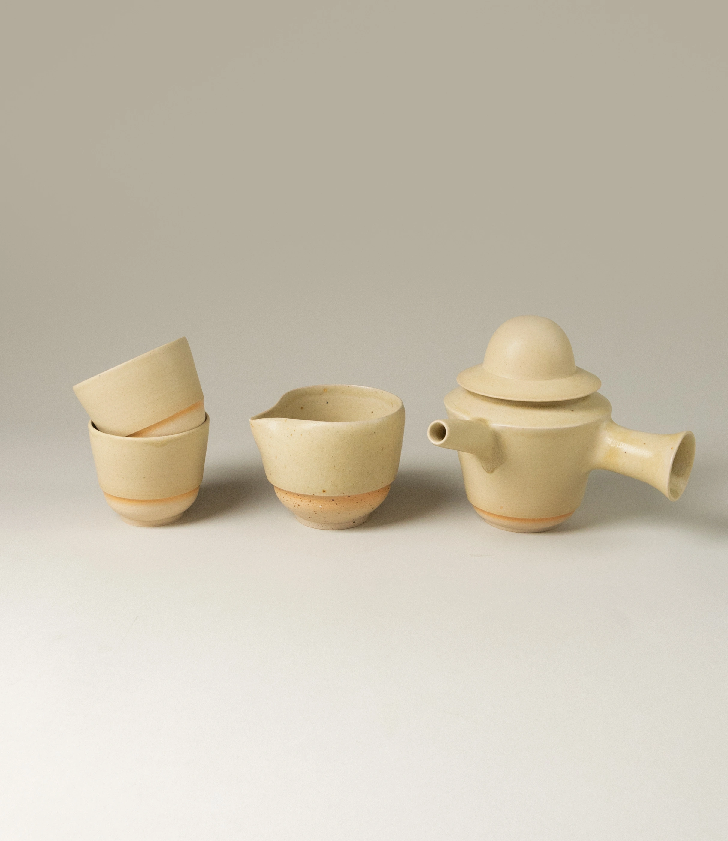 A clay tea set by Omni Ceramics, featuring a teapot with a unique spout design and lid on the right, a cream-colored cup with a pour spout in the center, and two stacked cups on the left, all displayed on a neutral background.