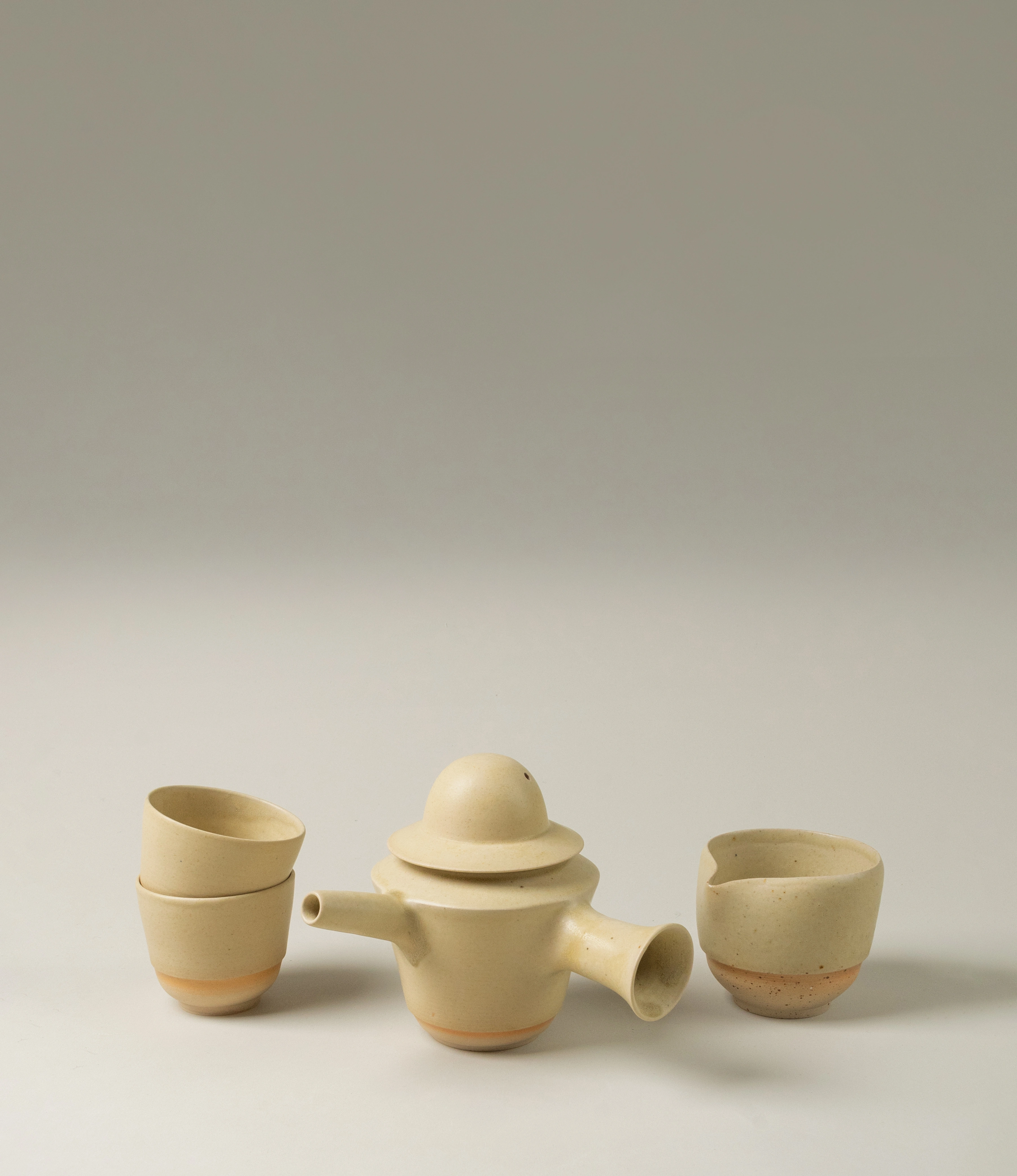 : "Omni Ceramics tea set displayed against a neutral background, featuring a clay teapot with a spout and lid, accompanied by two minimalist cups stacked on the left and one standalone pourer on the right.