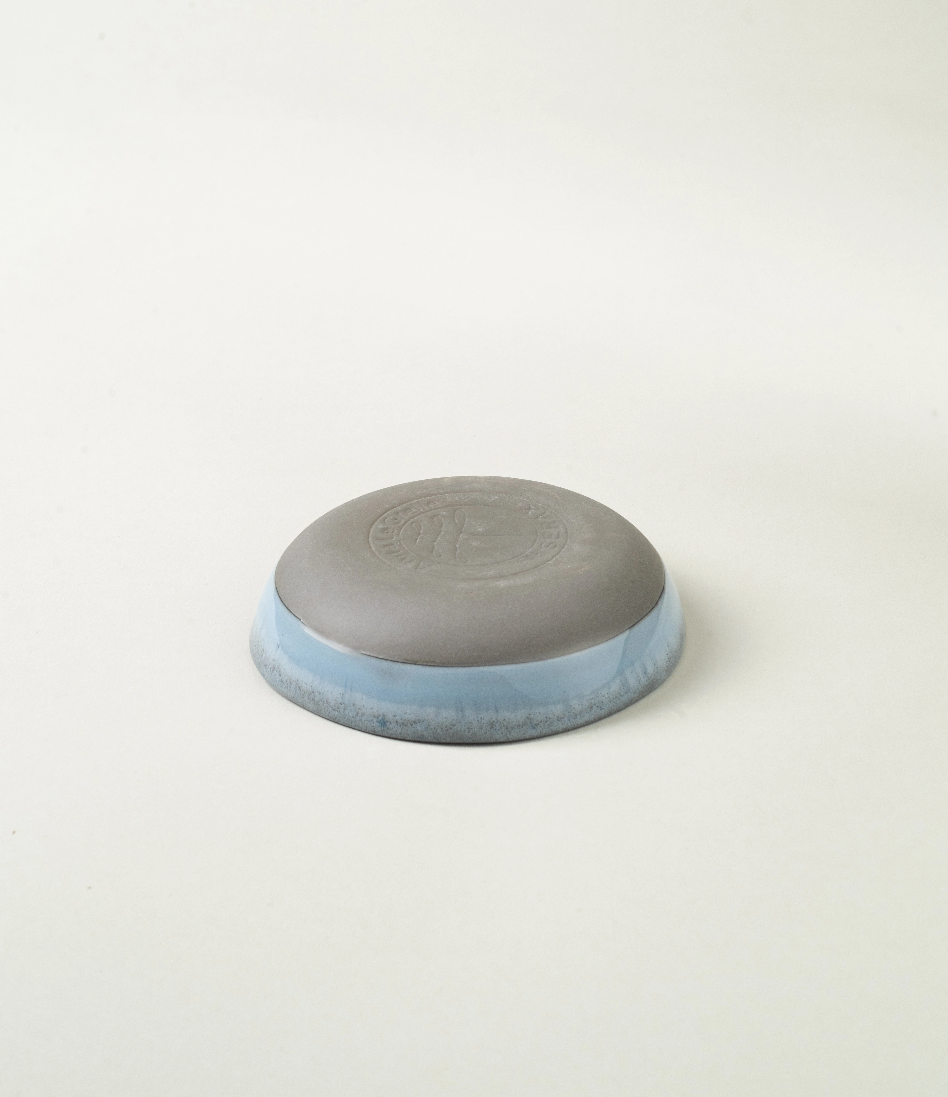 Blue Soap dish from Serax. The round item is made of ceramics and have a blue glazing with a grey unglazed matt area on the bottom. On the edges it has a grey rim. This picture shows the embossed bottom with the logo of Serax.