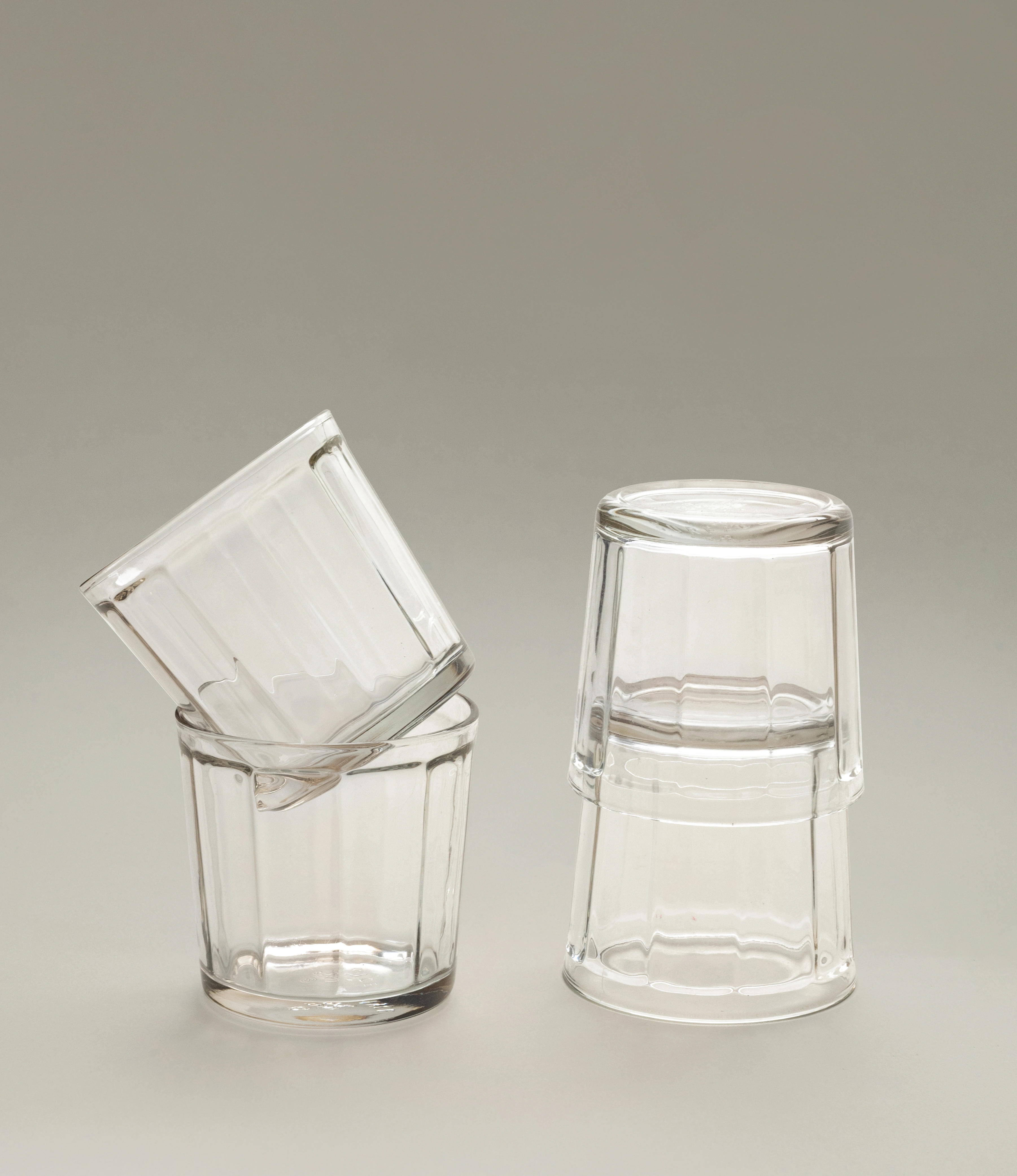 A set of clear glass tumblers by Serax, arranged with one glass tilted and nested inside another on the left, and two stacked upside-down on the right, showcased on a neutral background.