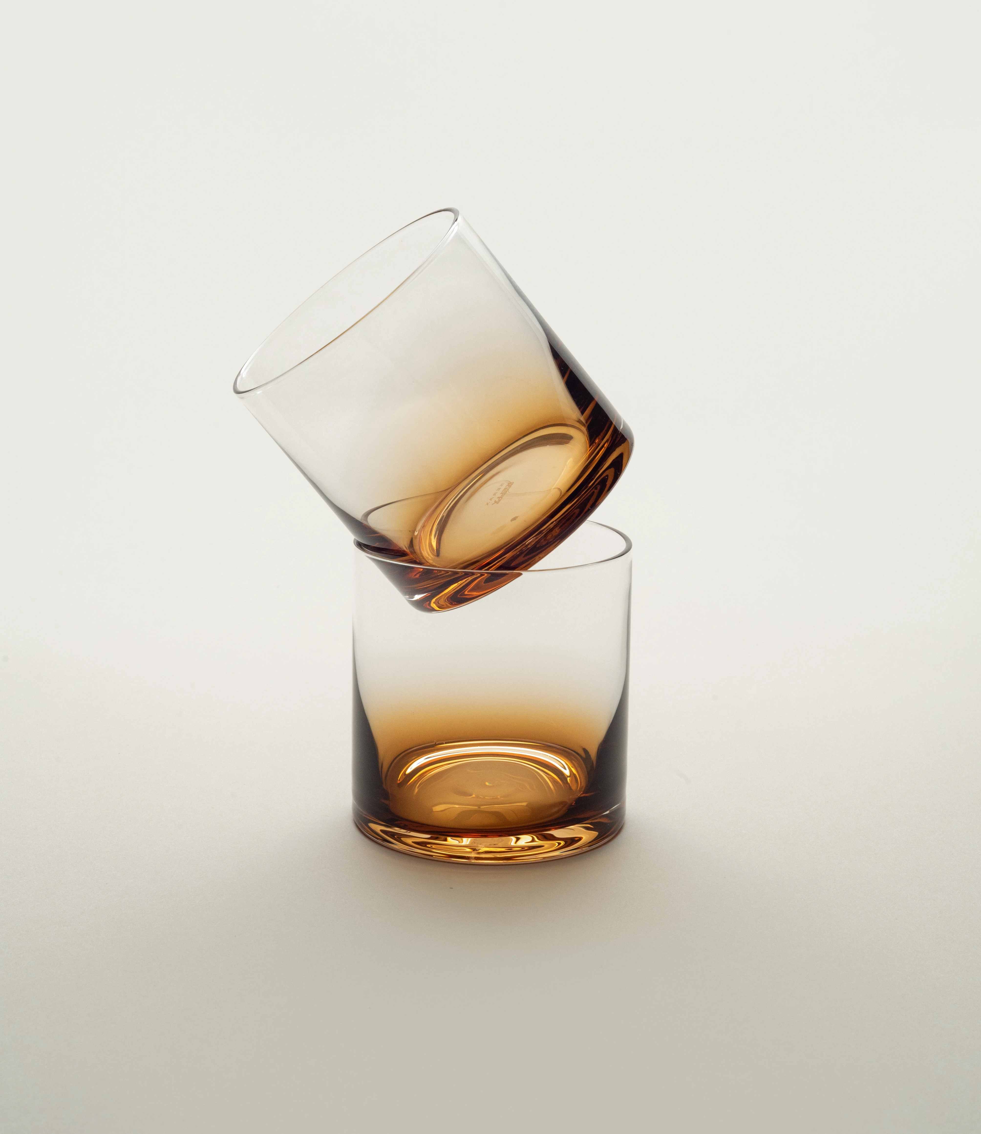 Two Serax Zuma whiskey glasses with amber gradient detail, one balancing atop the other against a minimalist background, showcasing the design's elegance and modernity.