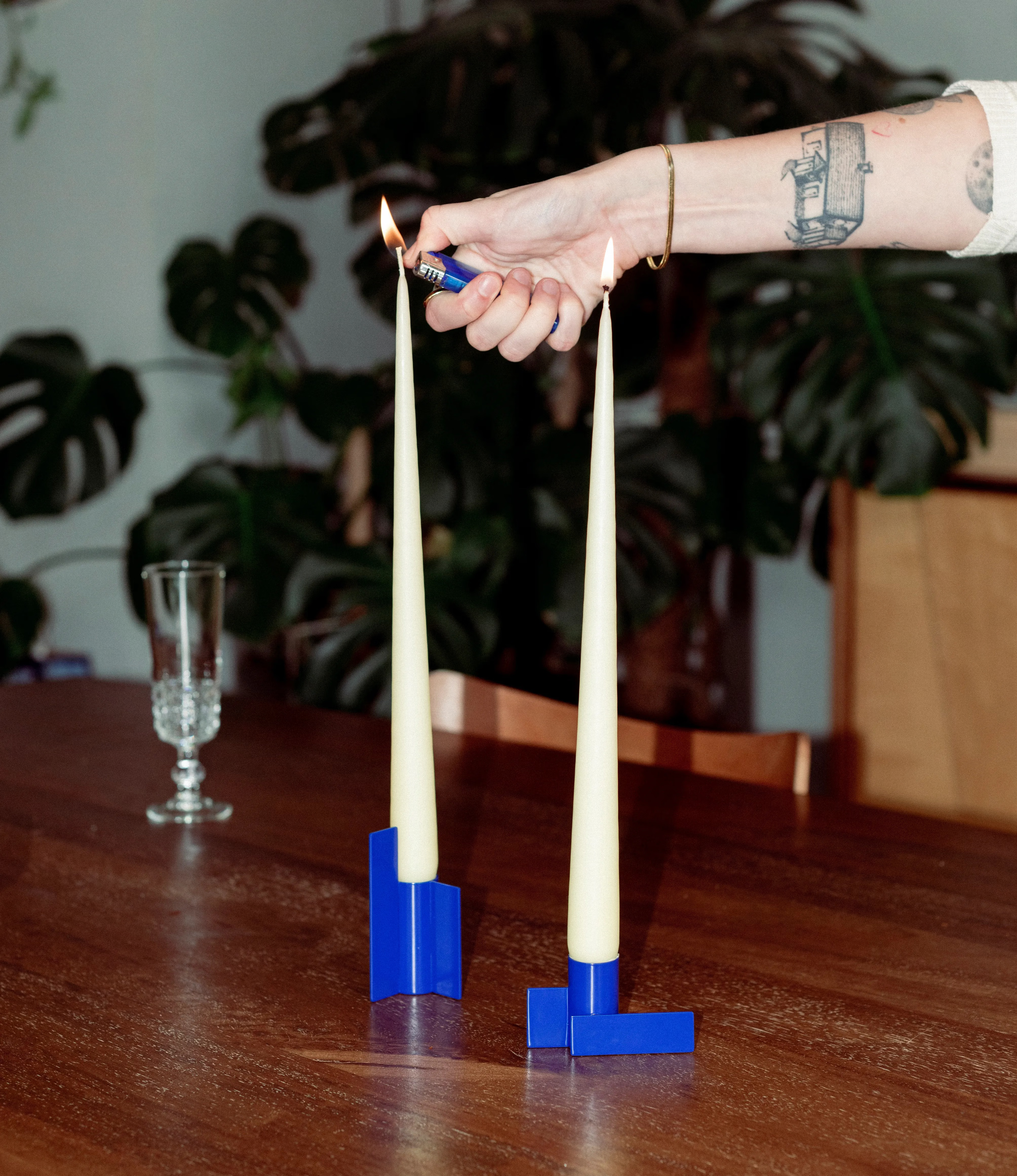Stences blue Icon Candle Holders are in use with the buttermilk taper candles. The candles are lit up by a hand on the picture.In the background there is a monsterra and a vintage glass for champagne.