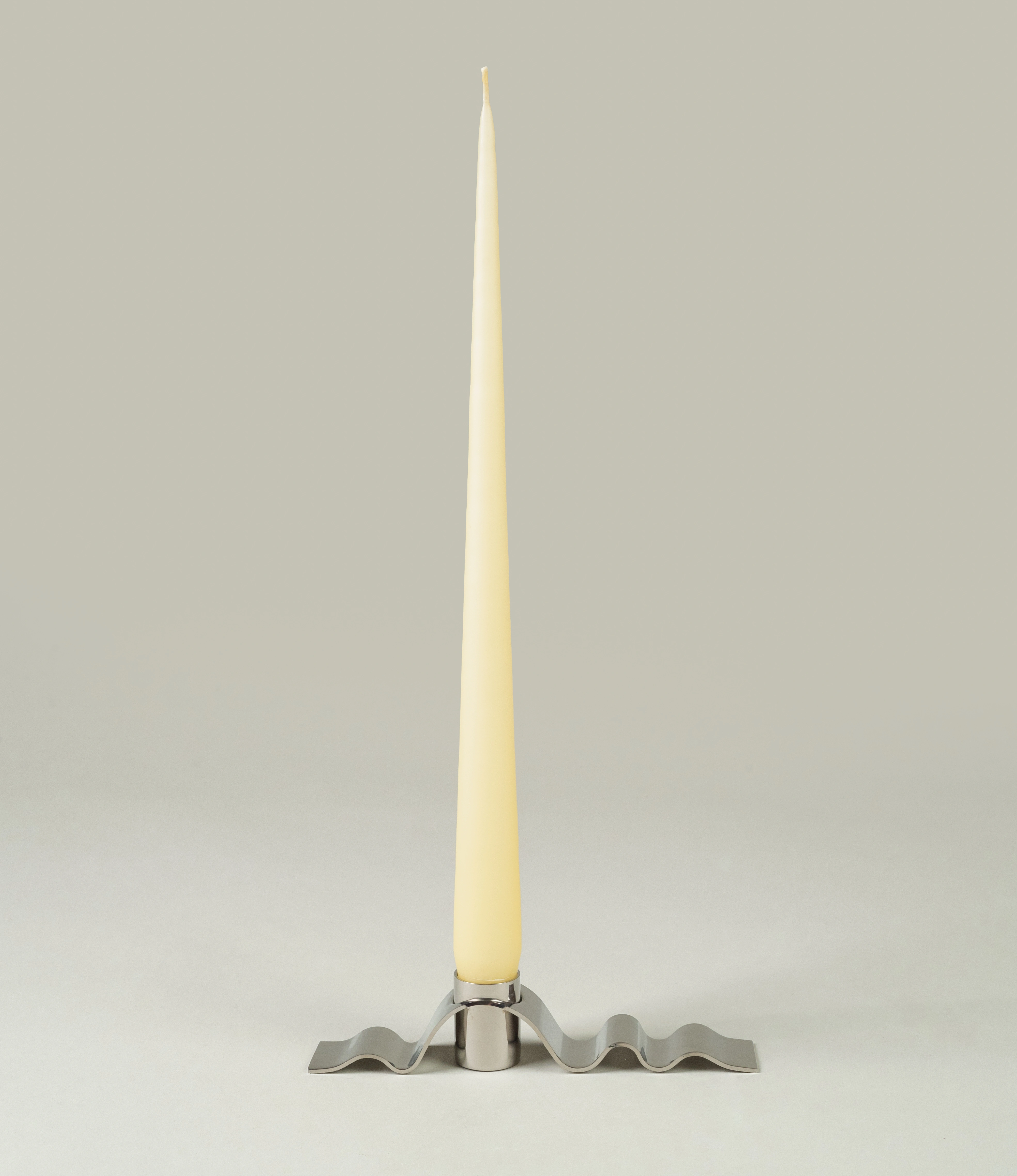 Wave candle holder in use with the taper candle from Ester and Eric coming in the buttermilk color. The candle is surrounded by a wavy stainless steel material which looks similar to a ribbon.