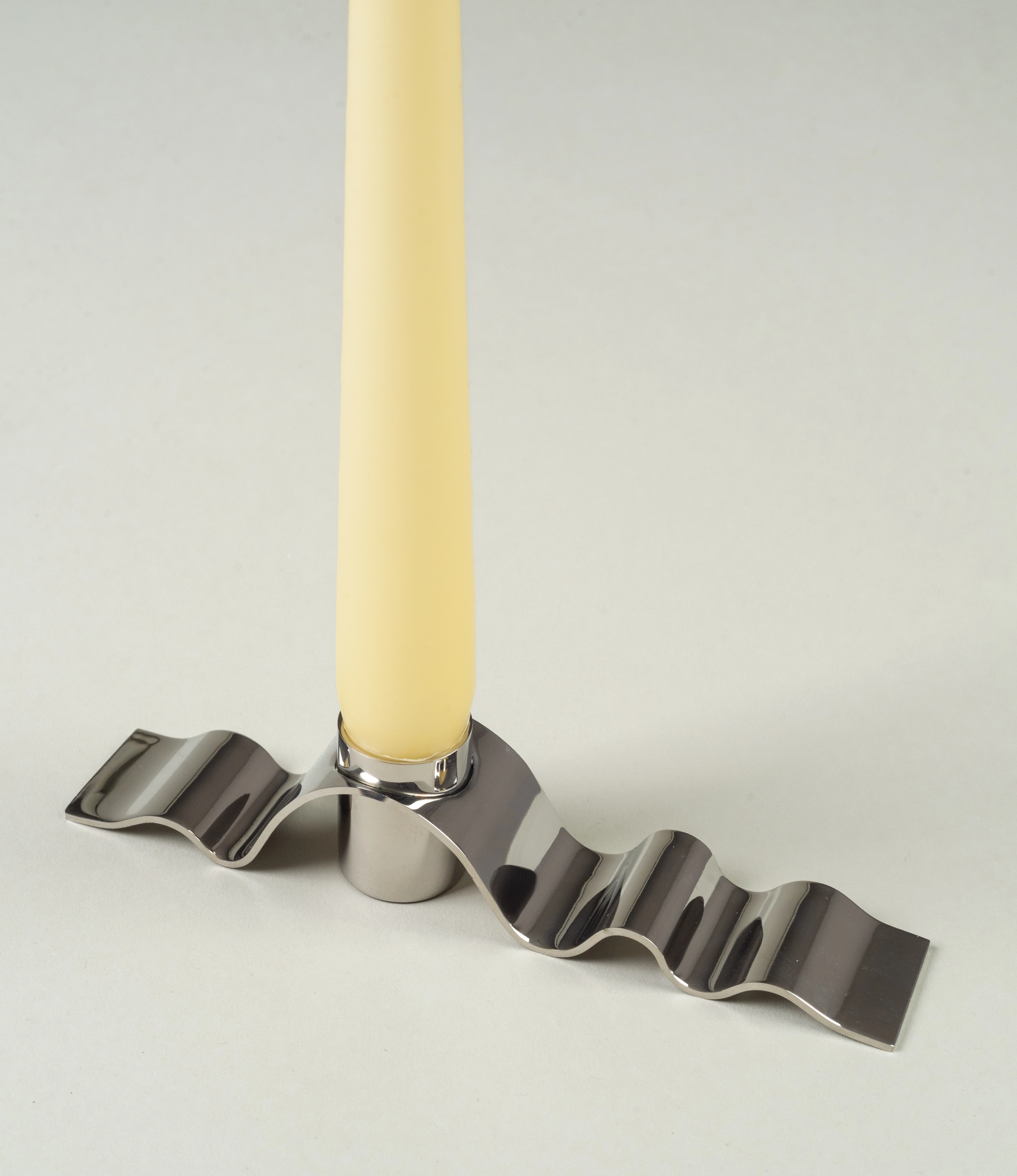 Wave candle holder in use with the taper candle from Ester and Eric coming in the buttermilk color. The candle is surrounded by a wavy stainless steel material which looks similar to a ribbon.