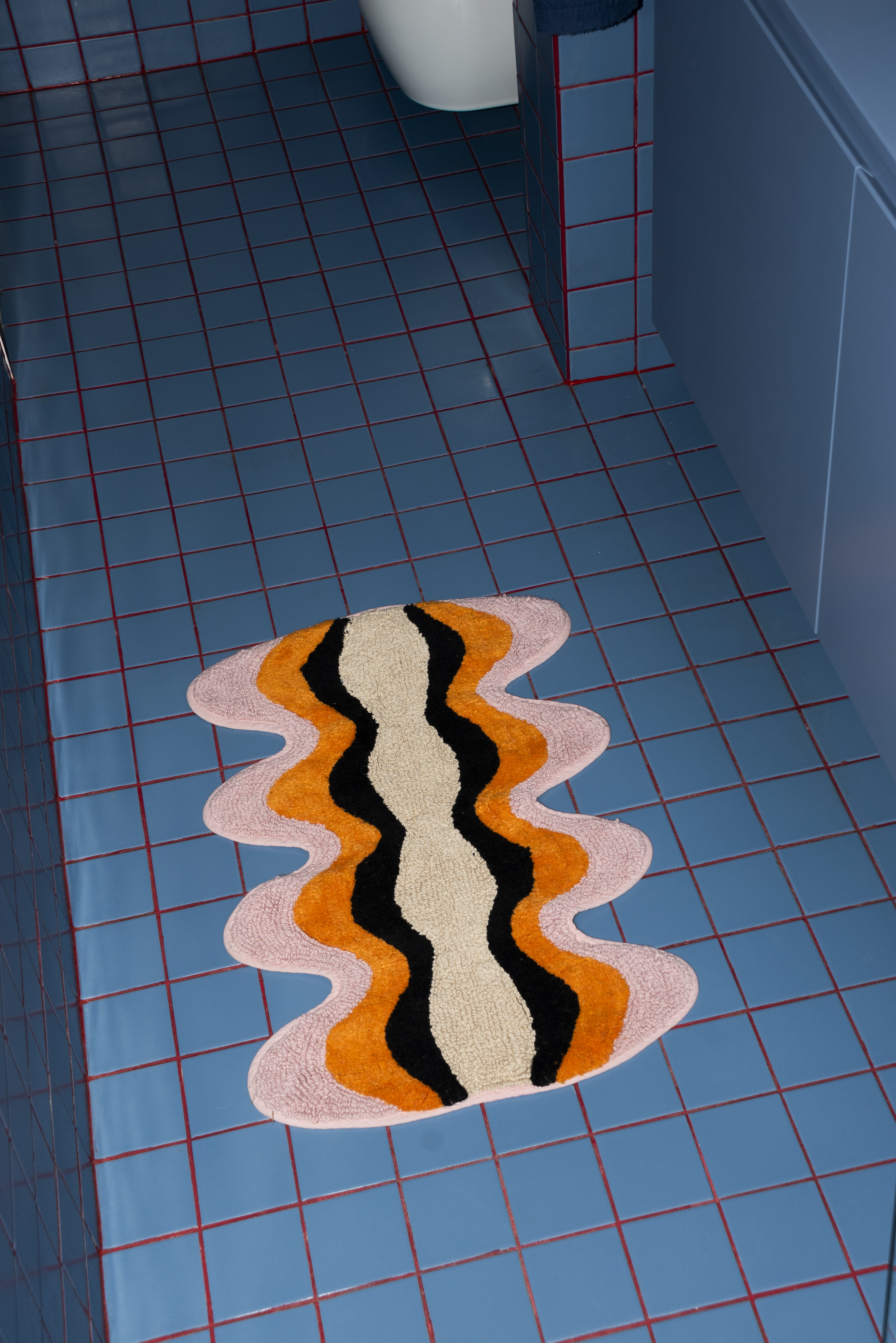 Tarta Gelatina Plankton Bathmat placed in a modern bathroom with blue tiled flooring, adding warmth and contrast with its bold, undulating pattern.