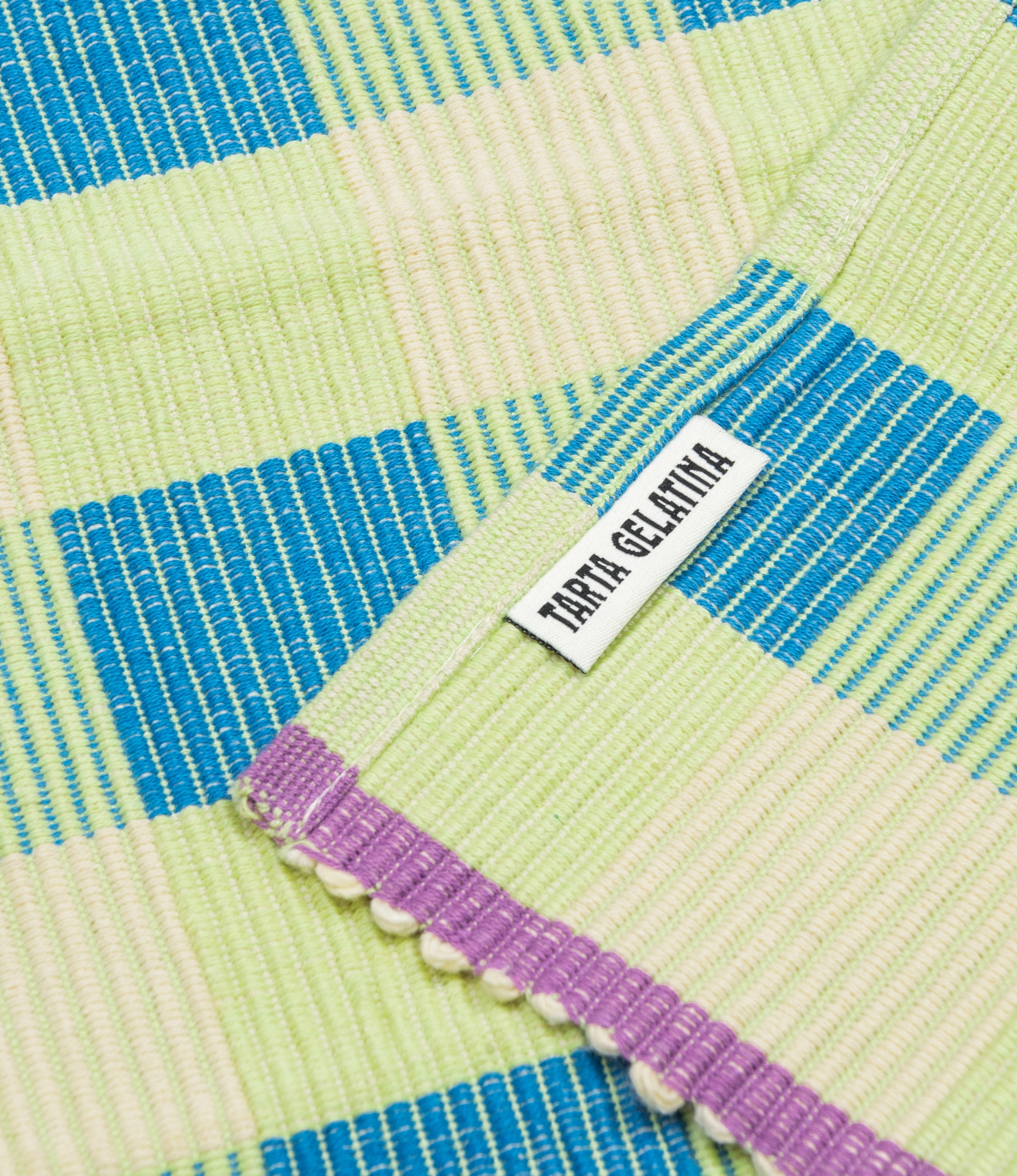 A closeup of Tarta Gelatina's Placemat coming in the green-blue variation. The edges are lilac on the shorter side. On this picture the logo of Tarta Gelatina is visible on small label sewed on the bottom of the product.
