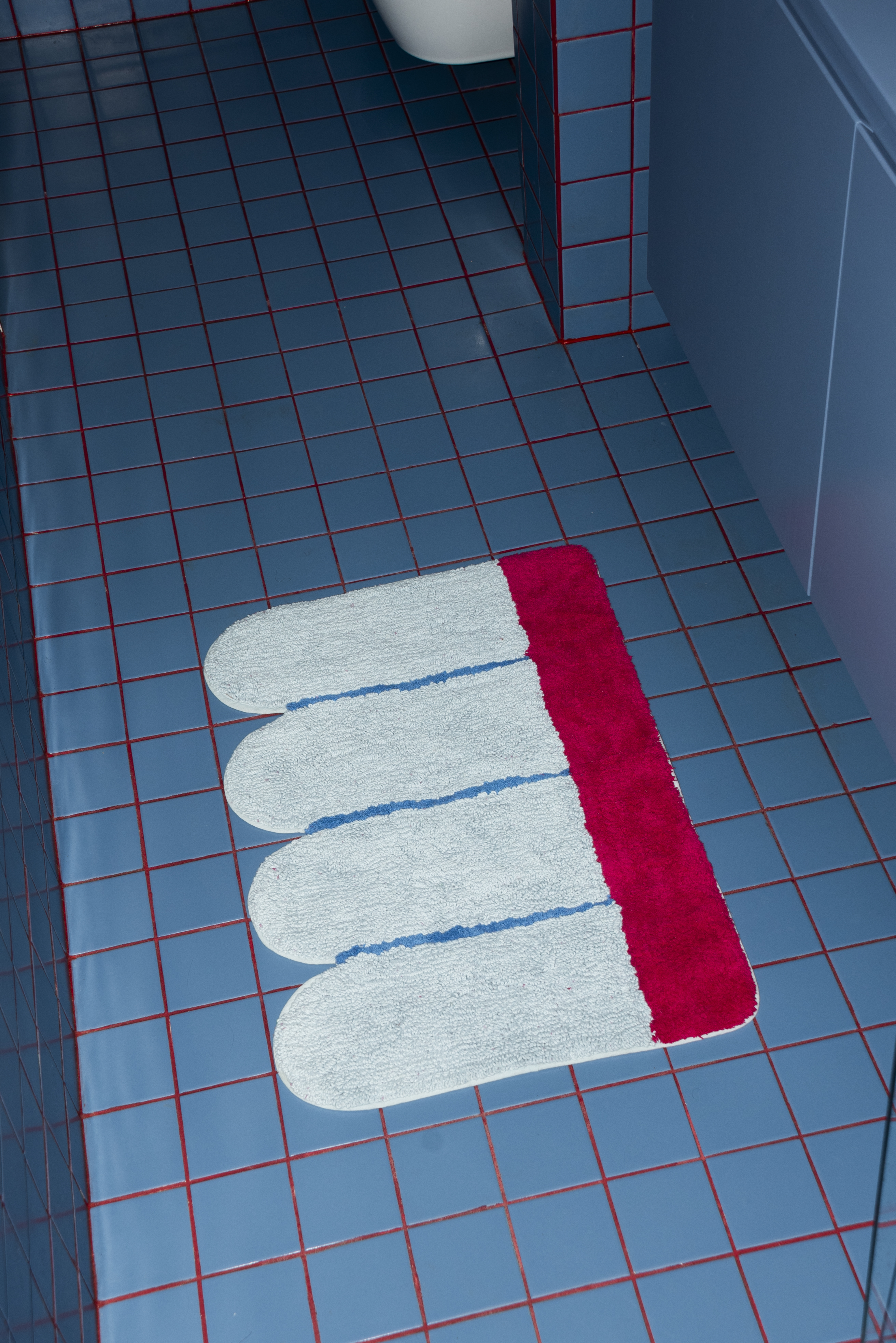 Close-up of the Poolside Bathmat By Tarta Gelatina placed on a blue tiled bathroom floor, highlighting its scalloped design and bold pink stripe.
