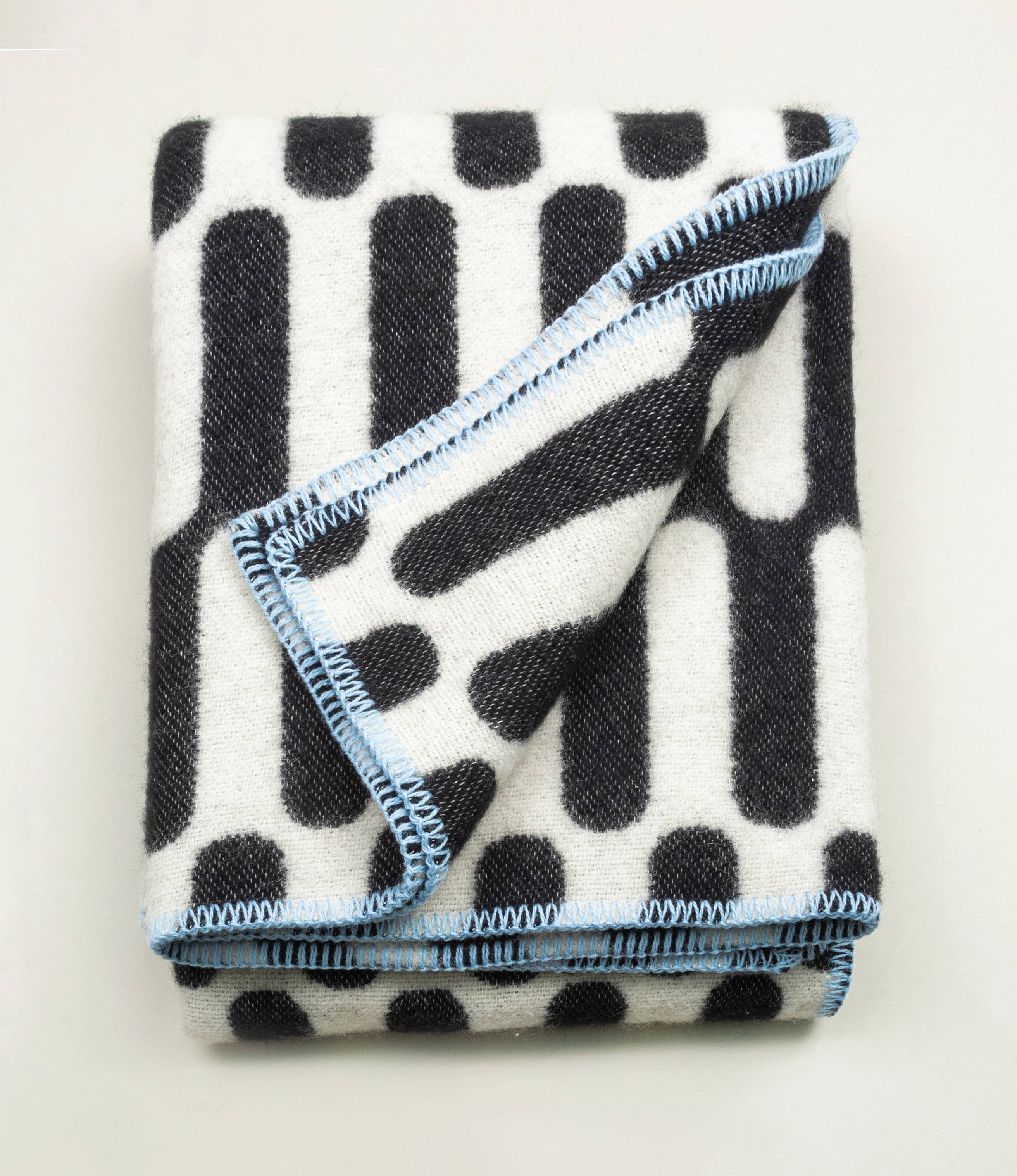 Throw Blanket From Tarta Gelatina coming in a black and white color. The pattern is similar to what you would see on op-art pictures. The edges are stiched with a light blue thread. In this picture the blanket is folded.