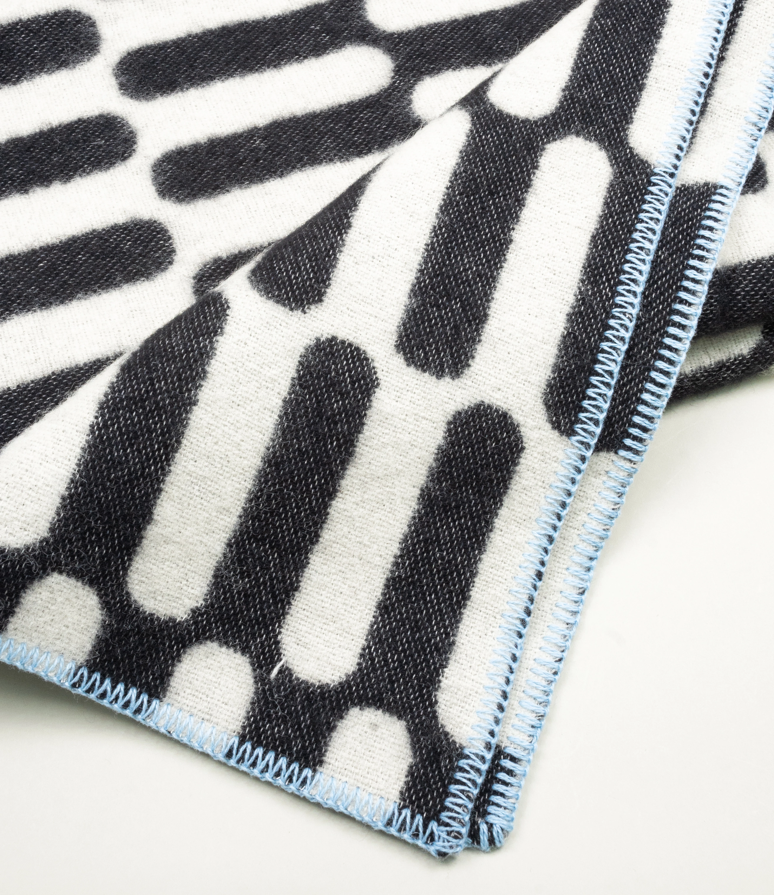 Throw Blanket From Tarta Gelatina coming in a black and white color. The pattern is similar to what you would see on op-art pictures. The edges are stiched with a light blue thread. In this picture the blanket shot from up close.