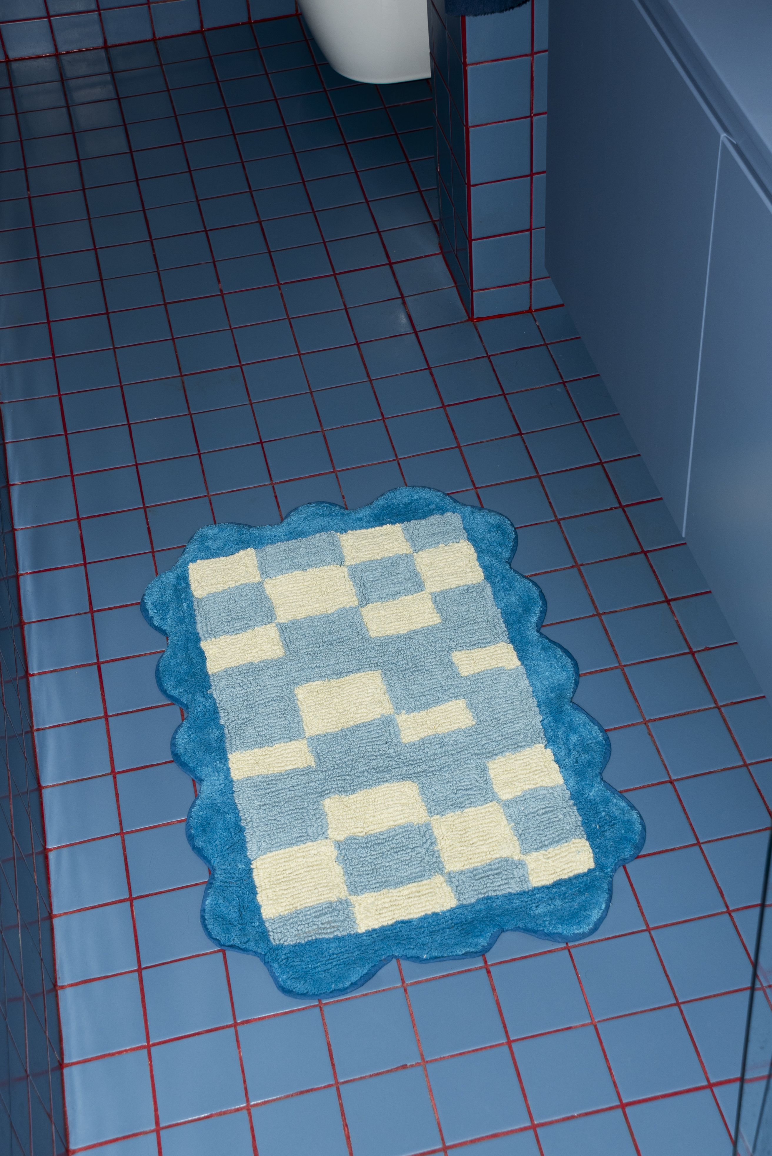 Tarta Gelatina's Burbuja Bathmat in a modern blue-tiled bathroom, featuring a soft scalloped edge and checkerboard pattern in blue and cream tones.