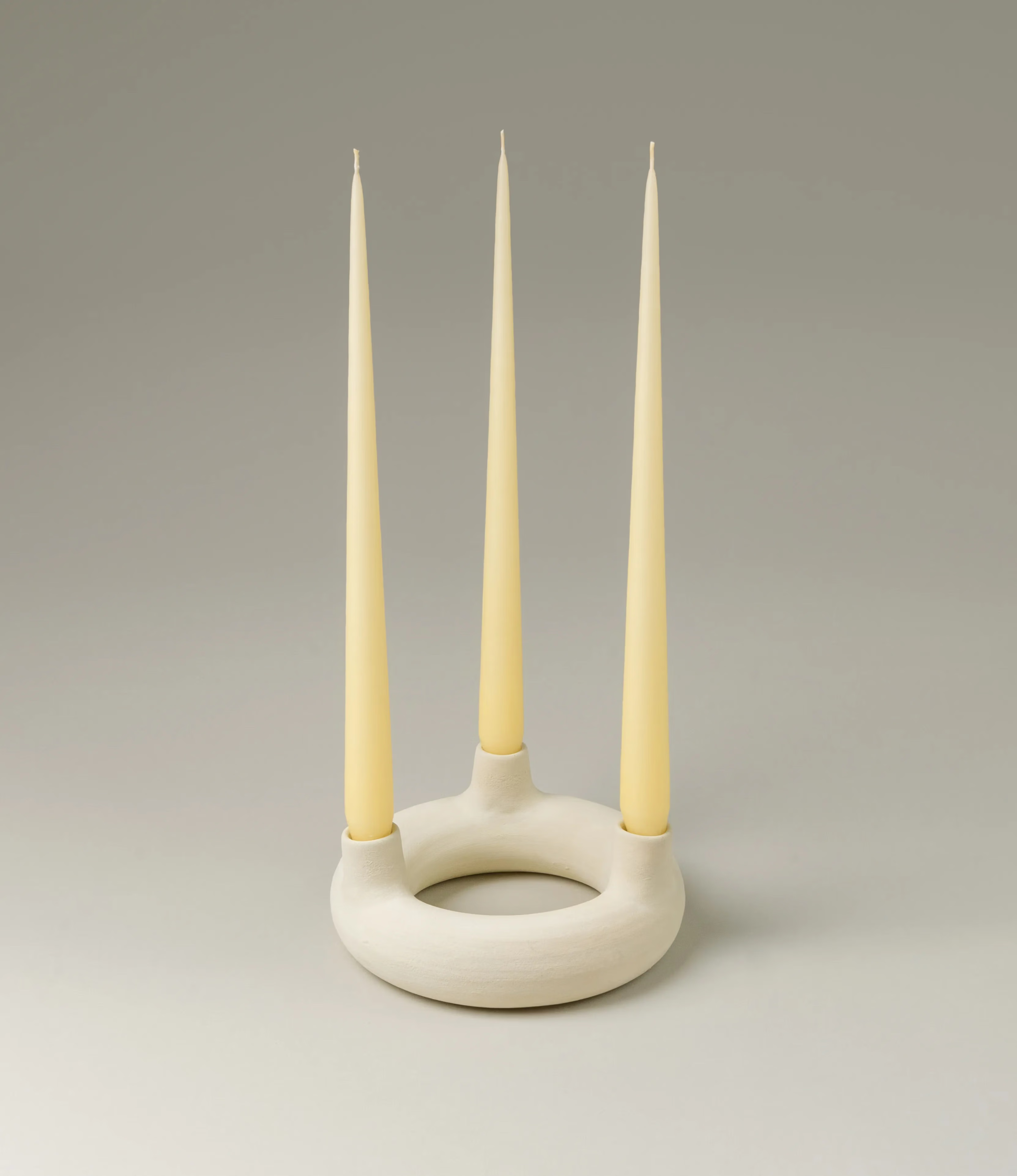 Chandelier Rotulis is a candle holder designed by Alfareria La Nava. The item can hold three candles arranged on a rounded shape. It has a matte raw white color. This picture shows the candleholder in use with three, light yellow colored taper candles from Ester&Erik