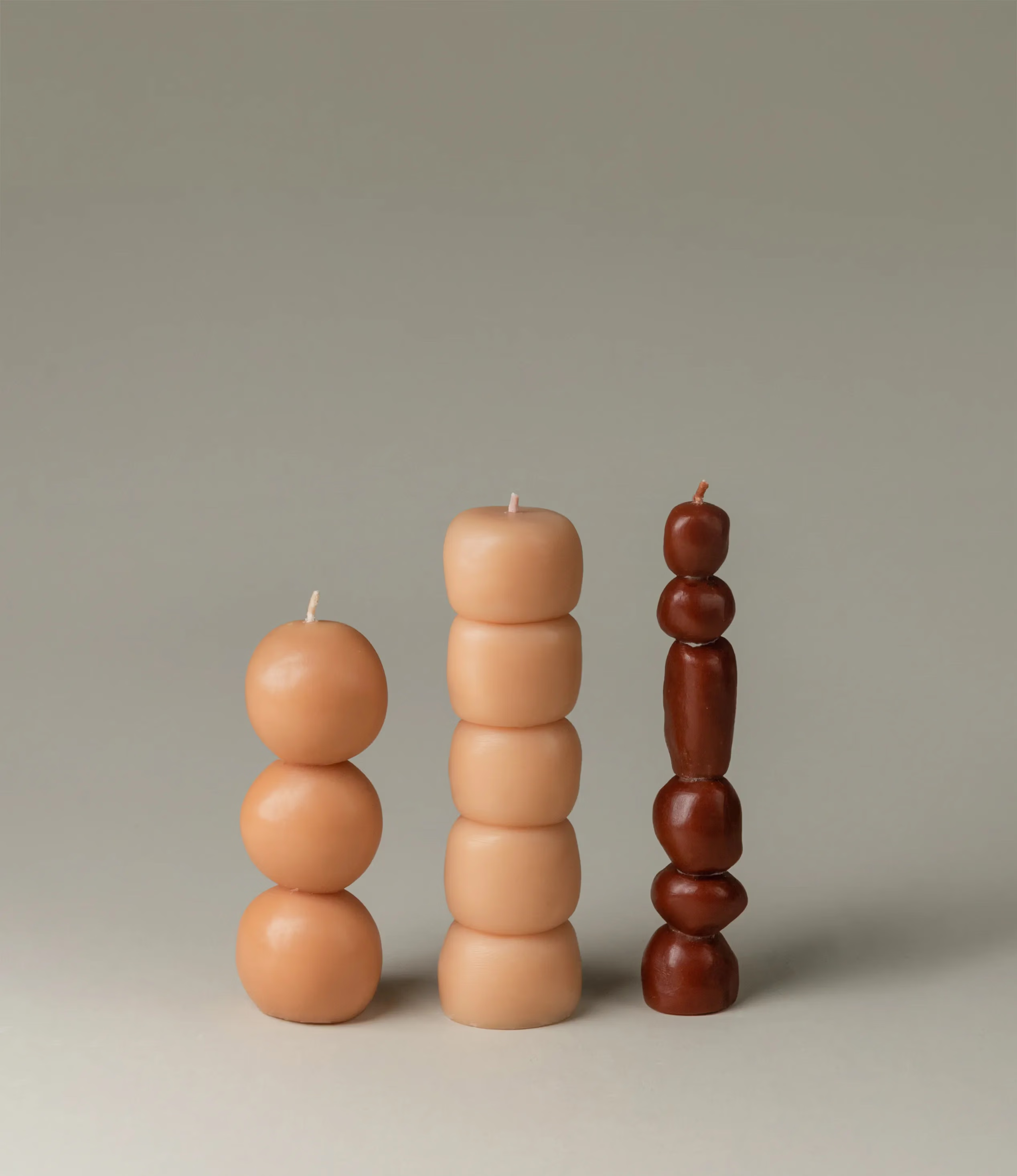 A packshot of the Piu set which contains three candles from Ann Vincent. One candle comes in a dark burgundy color the other two are beige. The candles look like little balls or stones stacked on top of each other.