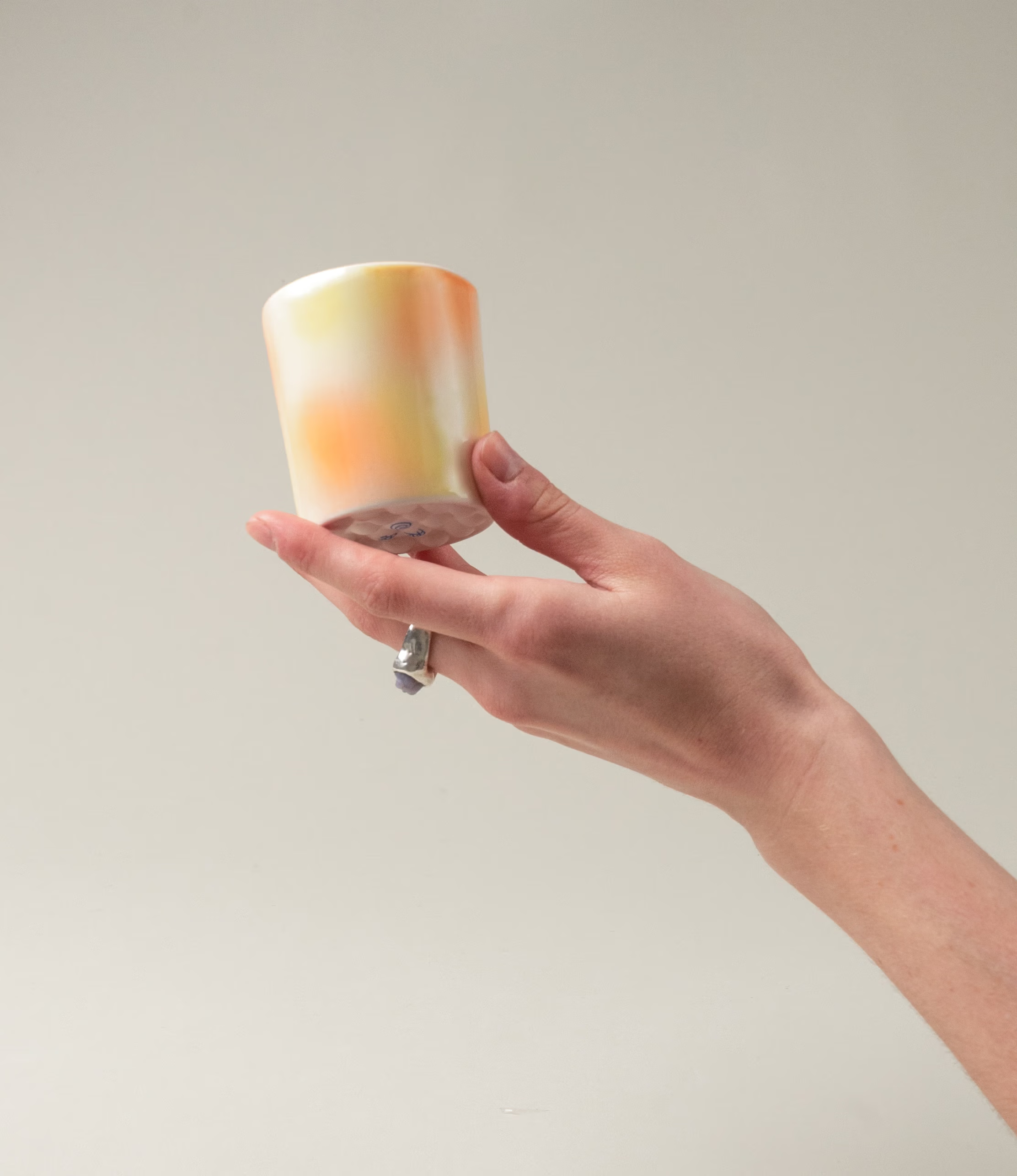 Bulle Cup from Frizbee Ceramics has a unique gradient painted pattern. The product has warm colors like orange, peach and yellow. The bottom of the cup looks like it's made of small bubbles. On this picture a hand is holding the cup.