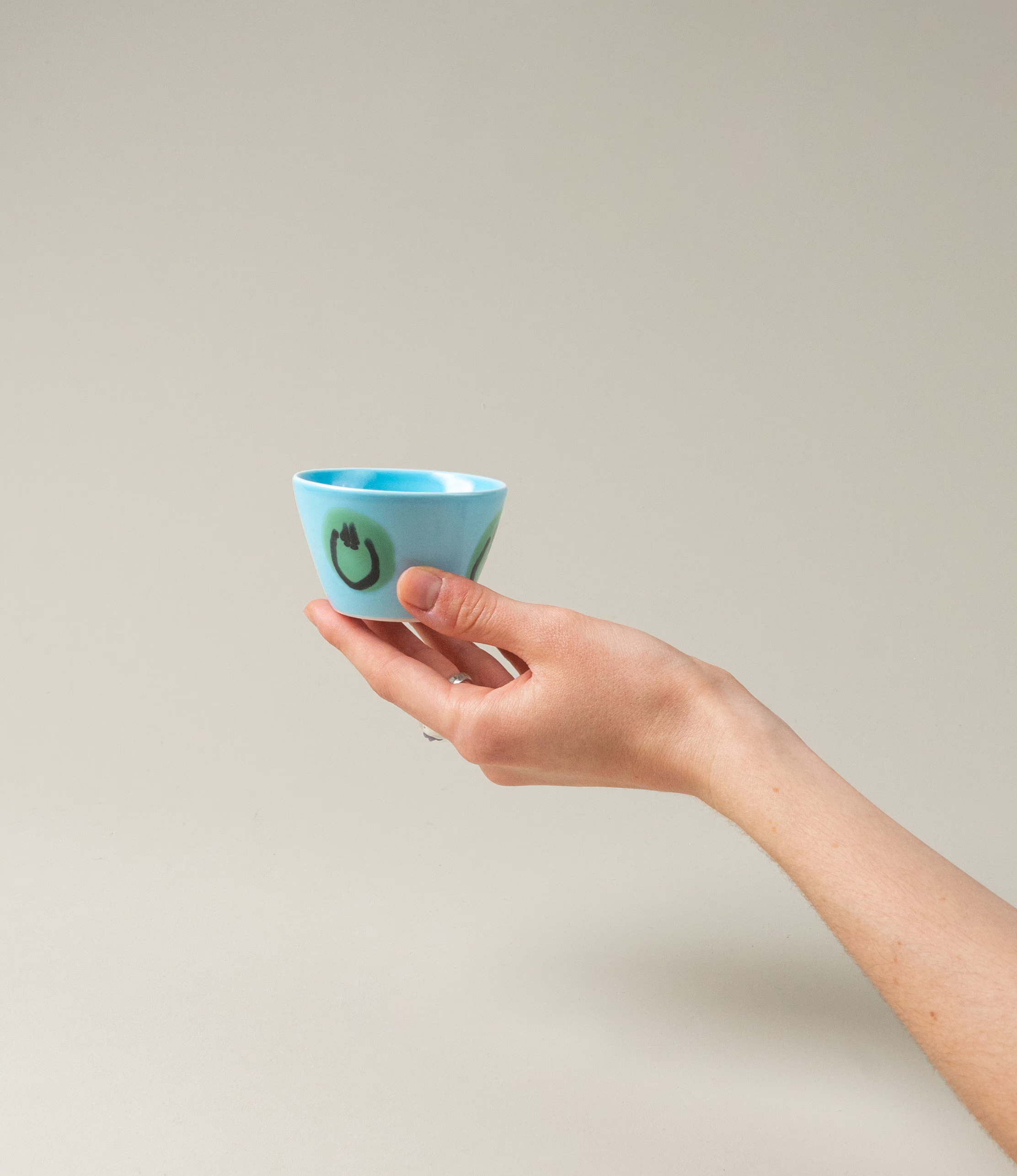 A hand holding the unique Blue Alien variant of the Super Cup Kawaii from Frizbee Ceramics. The item has green smiley pattern on a light blue base color.