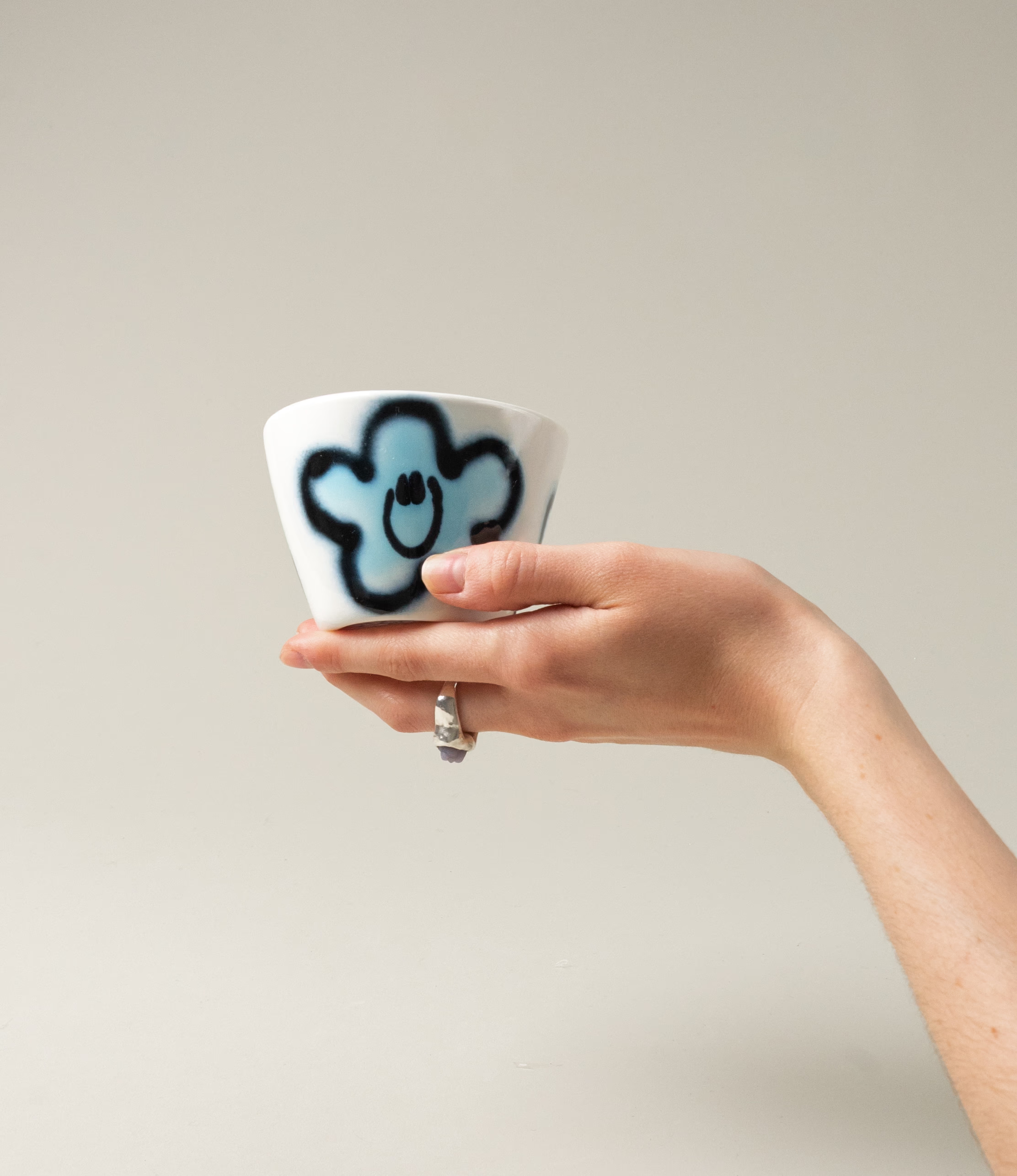 Super Cup from Frizbee Ceramics. The cup's base is white with a blue flower smiley. In this picture a hand is holding the cup.